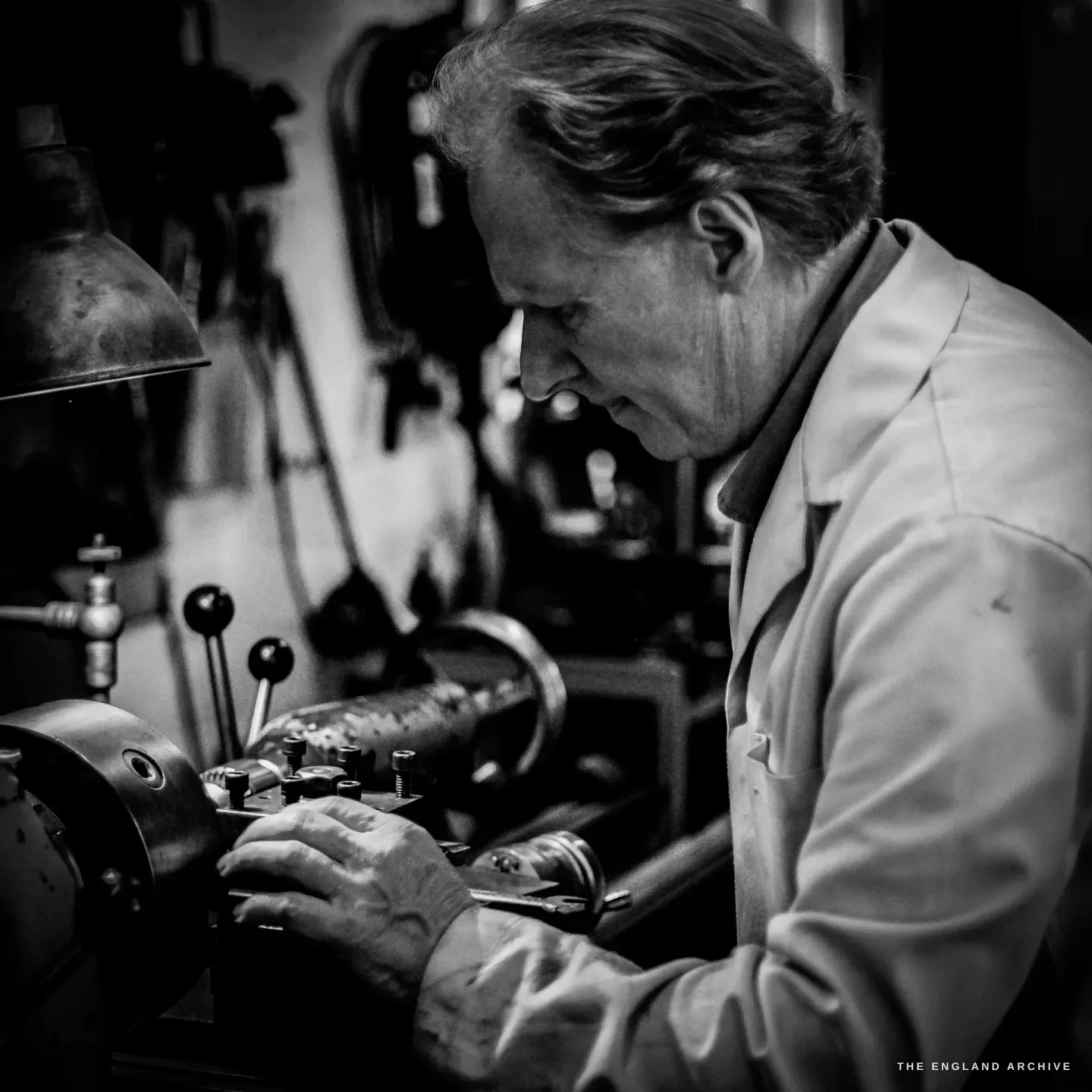 Haydn Hill in profile at an old lathe under an anglepoise lamp, in a pale work coat, warm light catching the brass and steel of the machine.