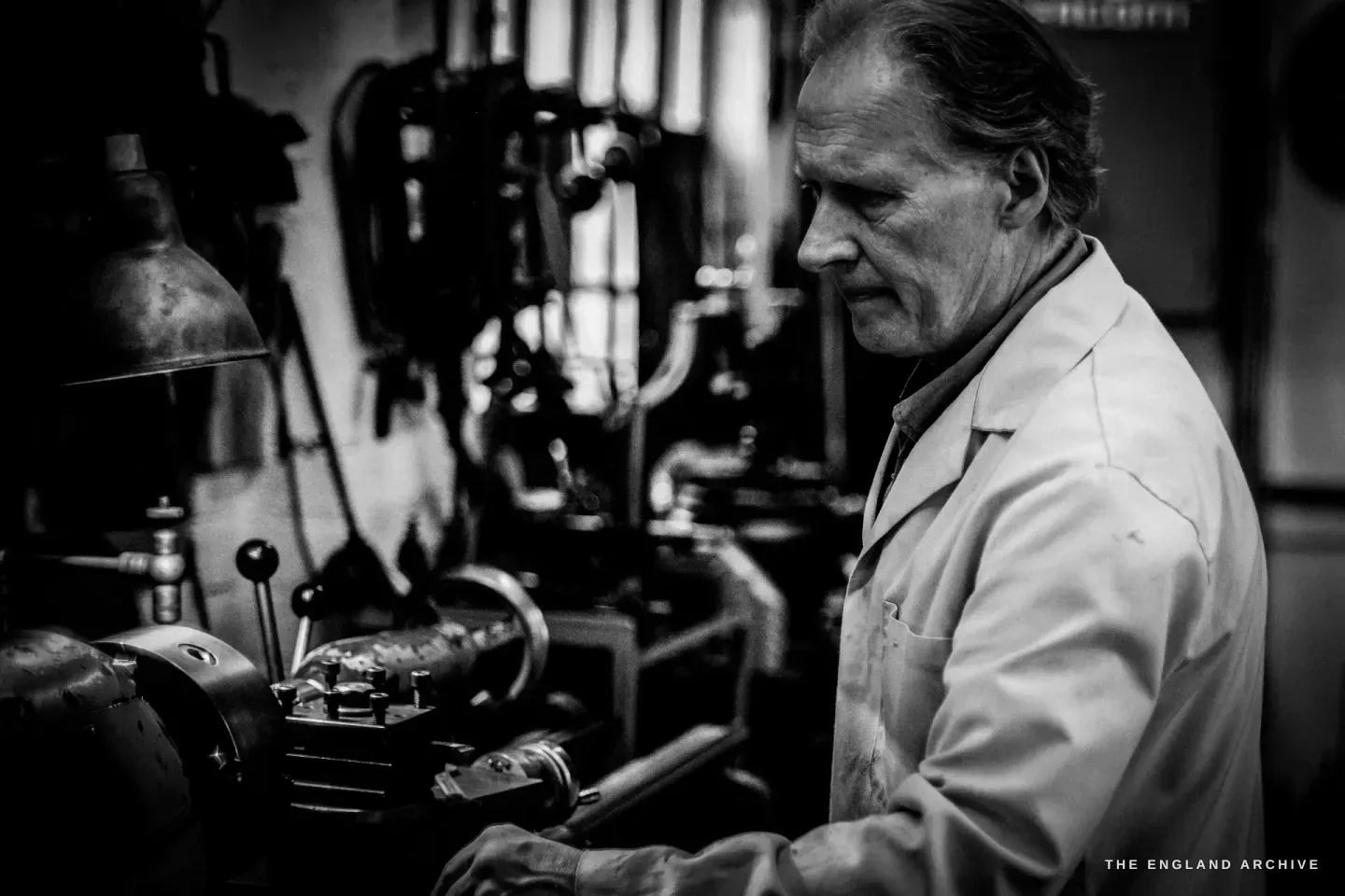 Haydn Hill at the lathe in three-quarter view, both hands on the work, a red cabinet and a wall of tools behind him in the dim workshop.