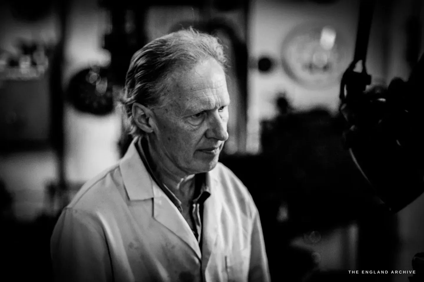 A close, low-lit portrait of Haydn Hill looking to one side in his white coat, the dark workshop behind him.