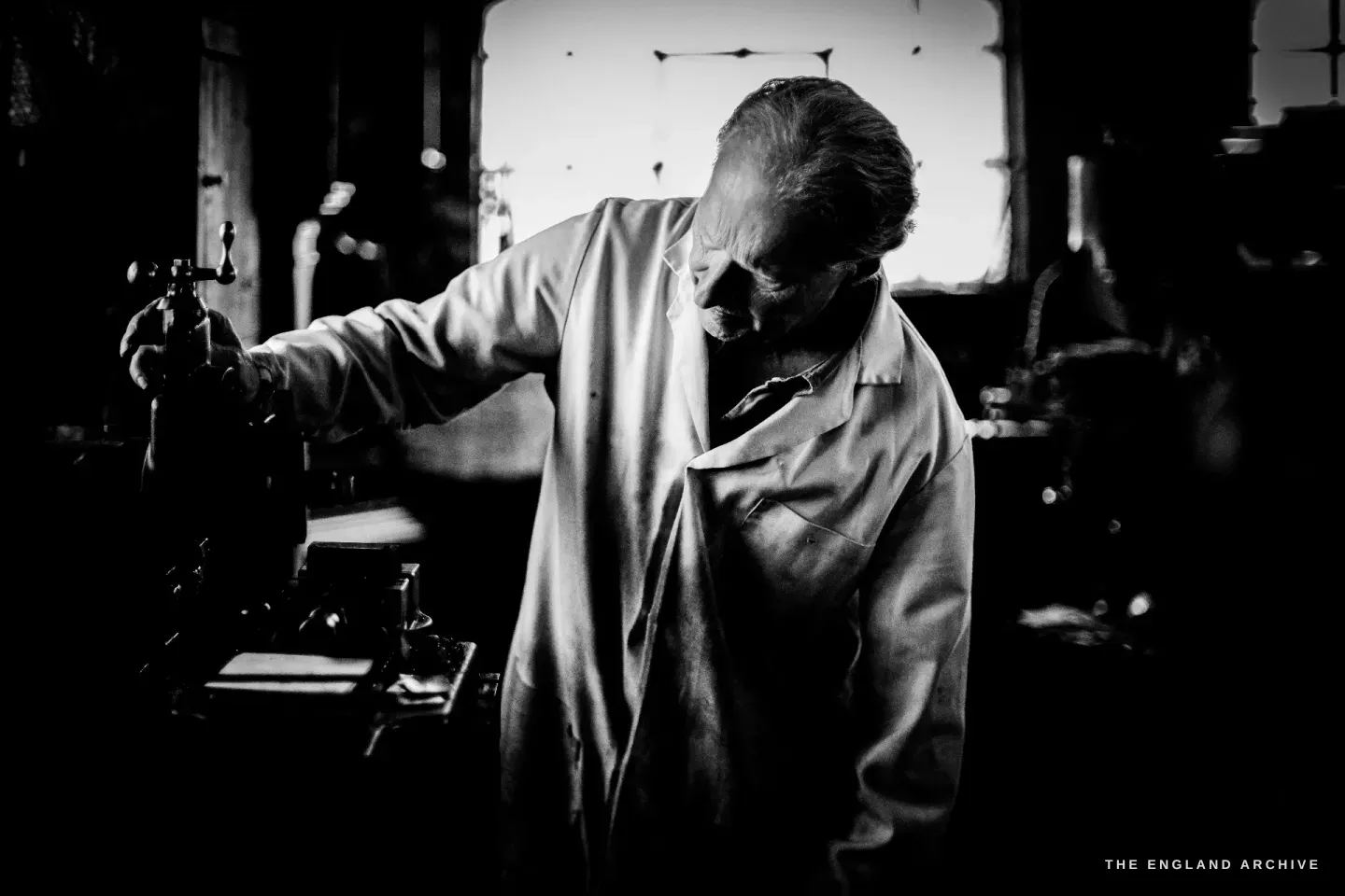 Haydn Hill bent low over a machine against a bright window, in silhouette and hard shadow, the workshop dark around the pool of light.