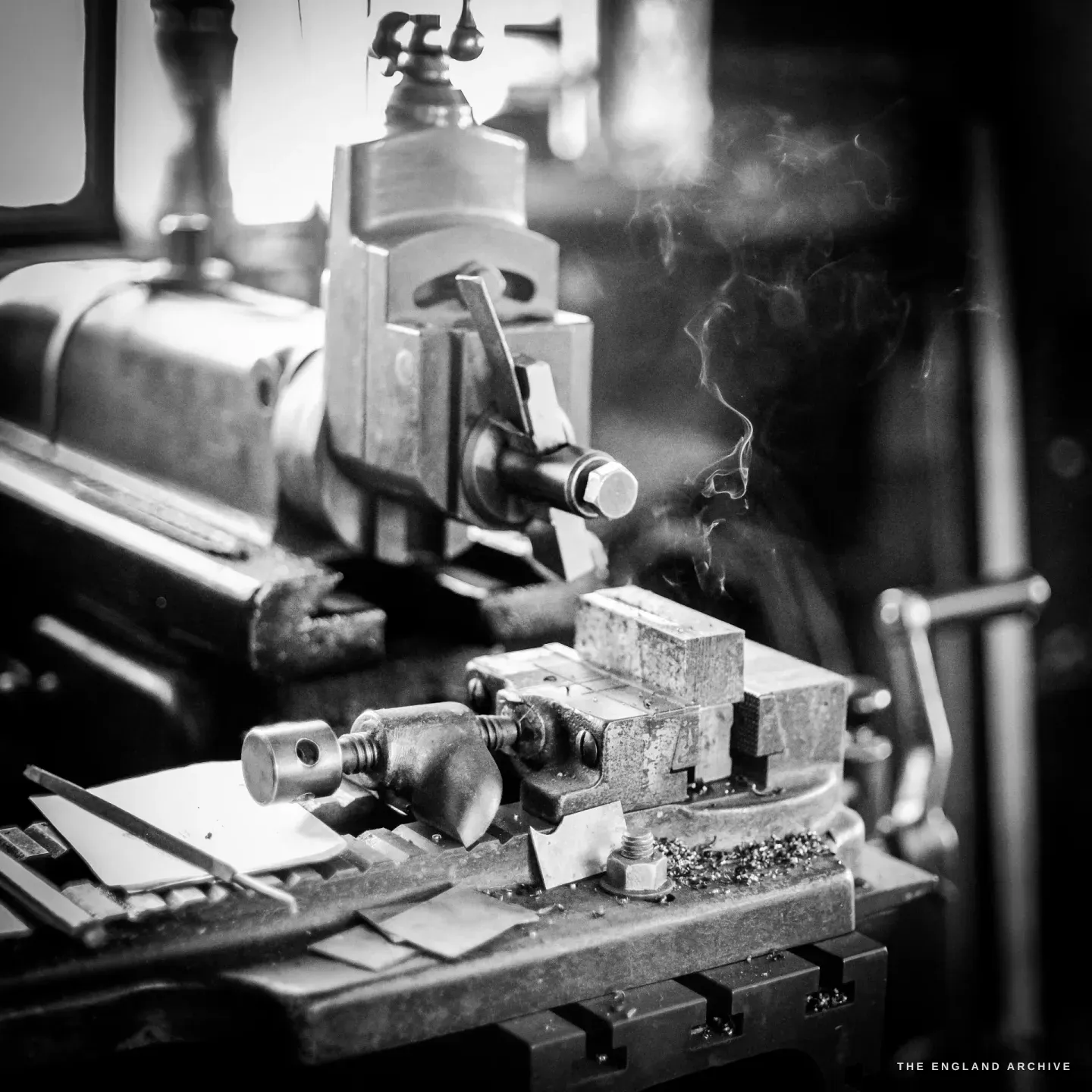 An old capstan lathe mid-cut, a thin curl of smoke lifting from the tool, bright swarf and filed flat parts on the machine bed, window light behind.