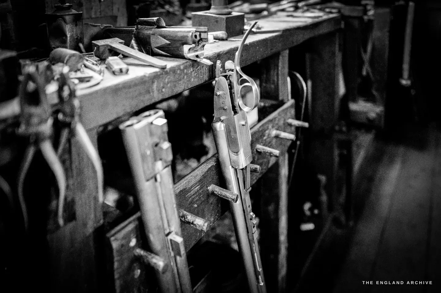 A shotgun action laid in a bench stand with its lockwork exposed, hand tools and a walnut forend on the bench beside it.