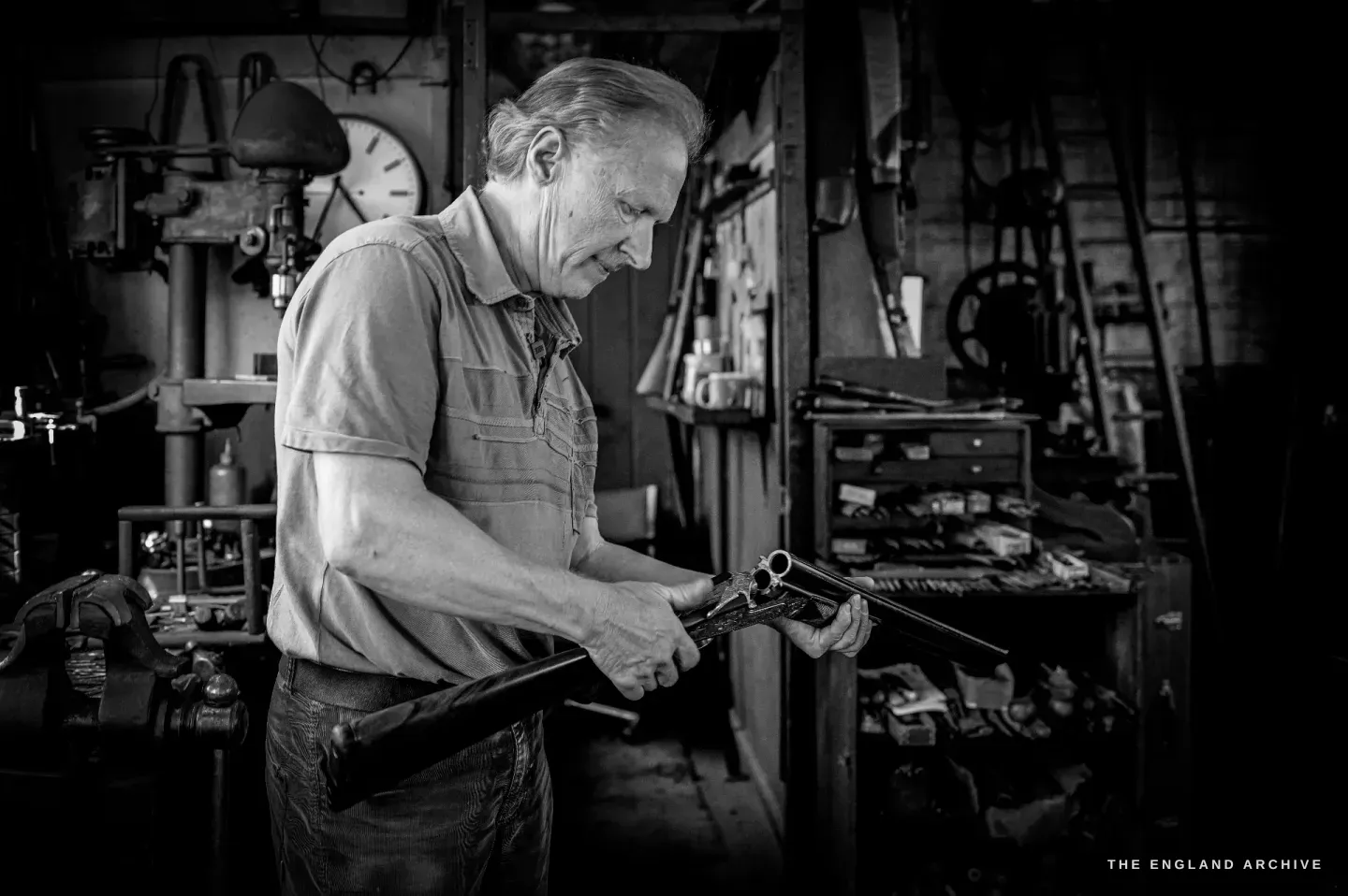 Haydn Hill stands at his bench turning a finished side-by-side shotgun in his hands, examining the barrels, a wall clock behind him.