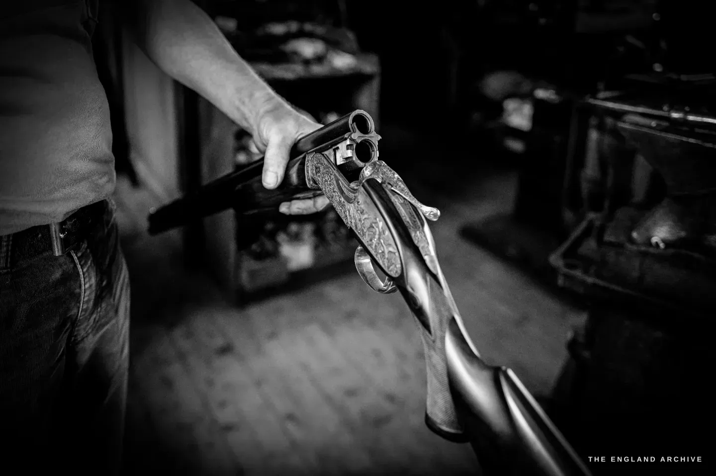 A finished side-by-side gun broken open in the hands, the scroll-engraved action and the deep-figured walnut stock catching the light.