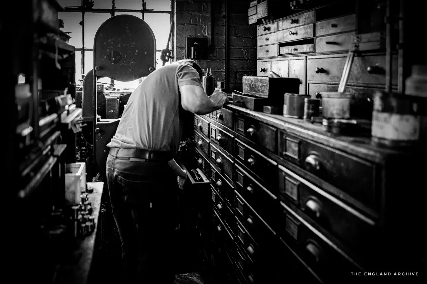 Haydn Hill seen from behind, reaching into one of a bank of small black tool drawers by a bright window, a band saw beside him.