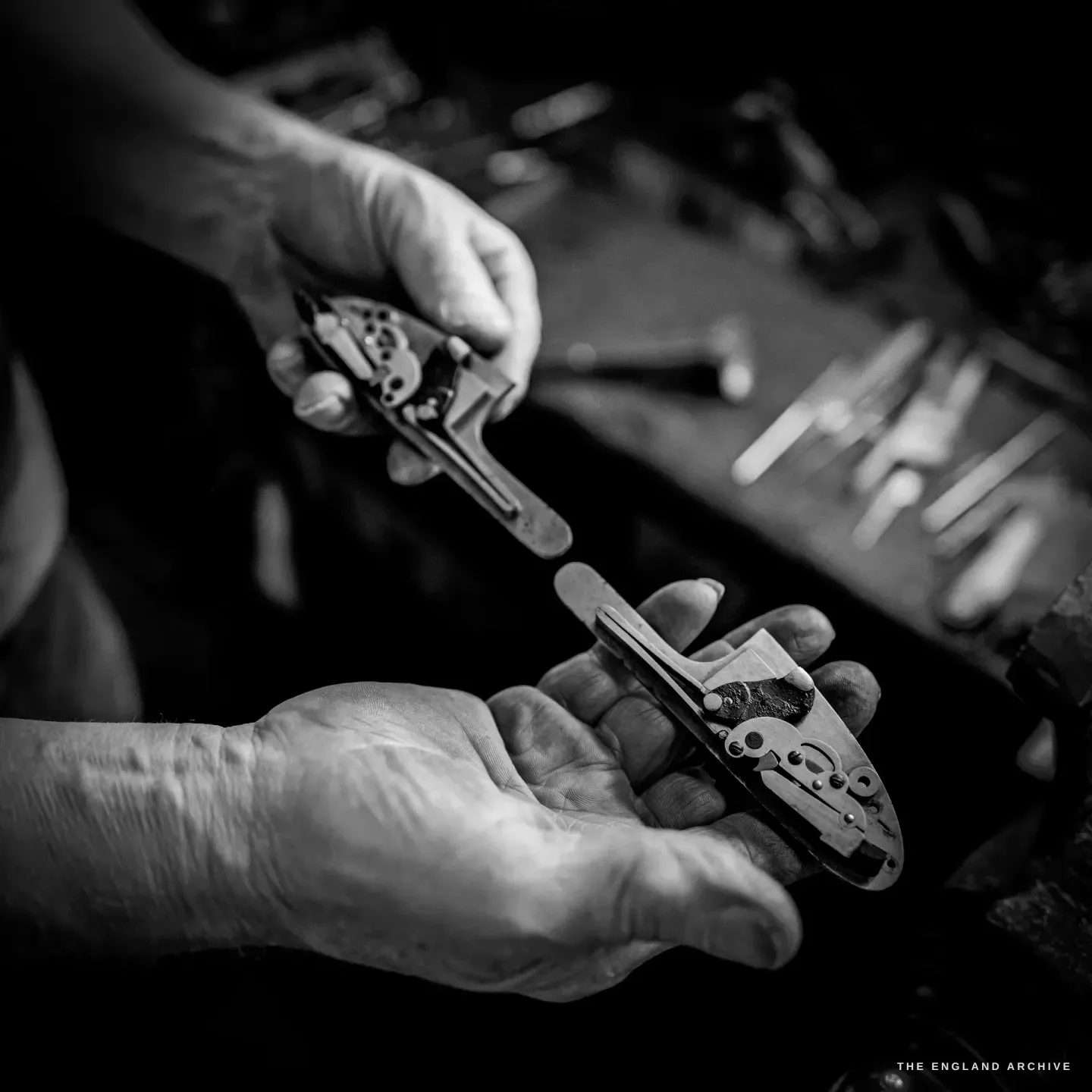 Two hands each hold a sidelock plate up for comparison, the fine lockwork of each visible against the dark workshop.