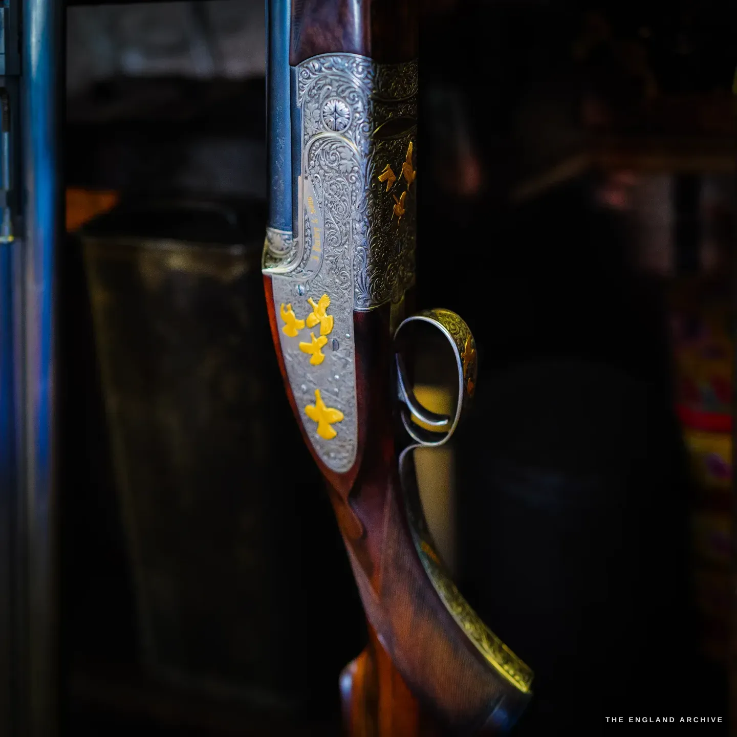 A close view of a finished best gun: fine scroll engraving across the action inlaid with gold game birds in flight, the stock deep-figured walnut, a blue tin beyond.