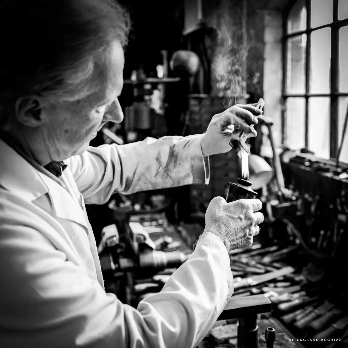 Haydn Hill holds a small steel component in the flame of a spirit lamp by a tall window, watching the colour of the metal, tools racked below.
