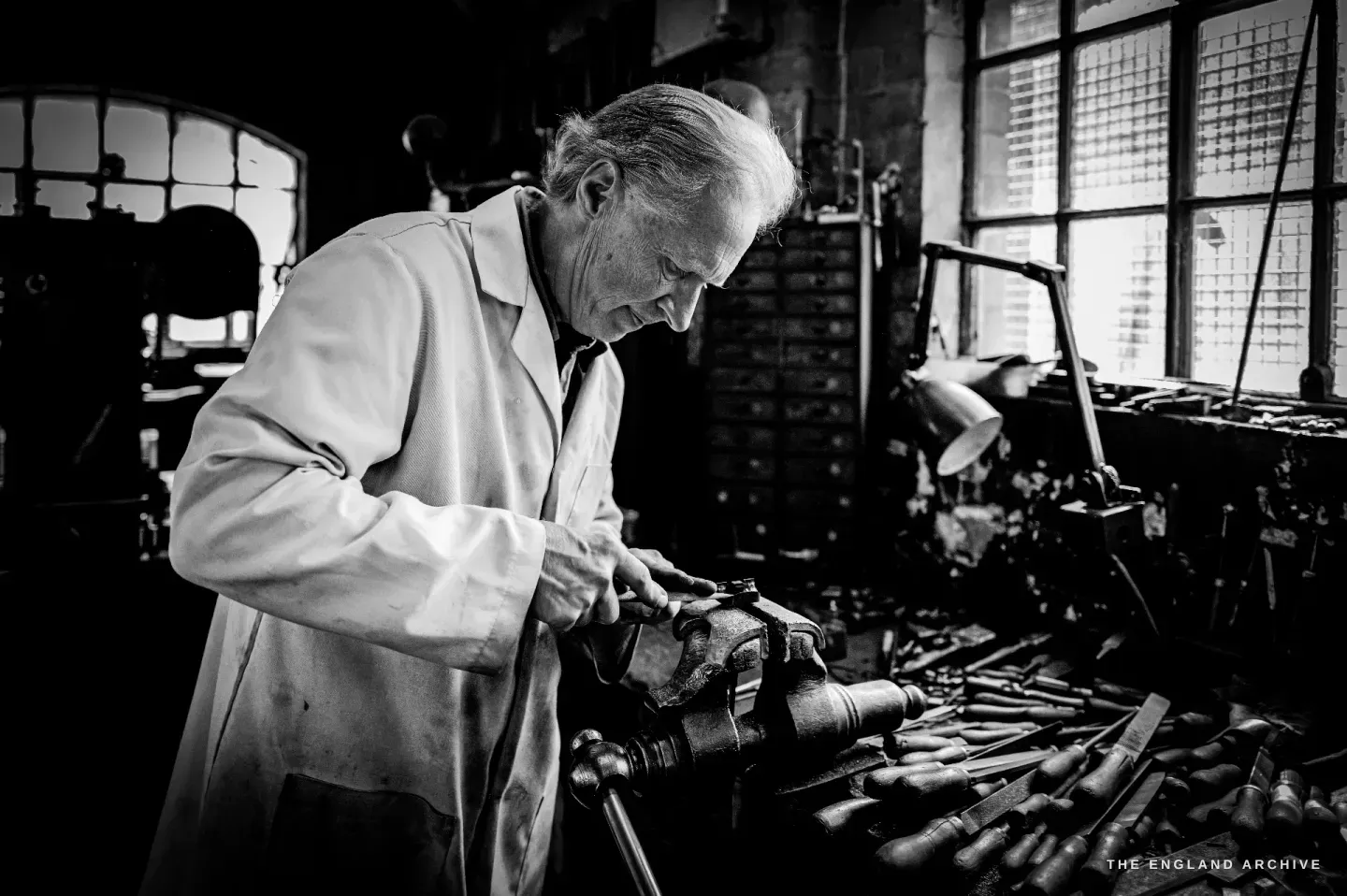 Haydn Hill at the bench working a part in a machine, a wide daylit view of the workshop with an arched window and a bench thick with tools.