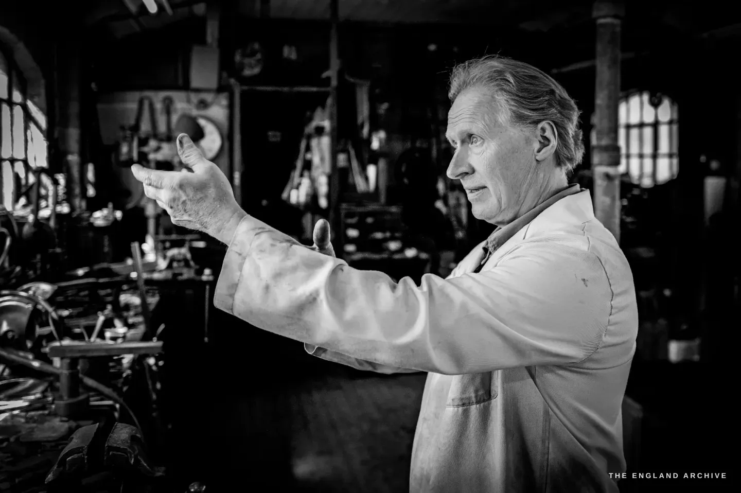 Haydn Hill in profile in his white coat, one arm outstretched as he explains something, the machines of the workshop around him.