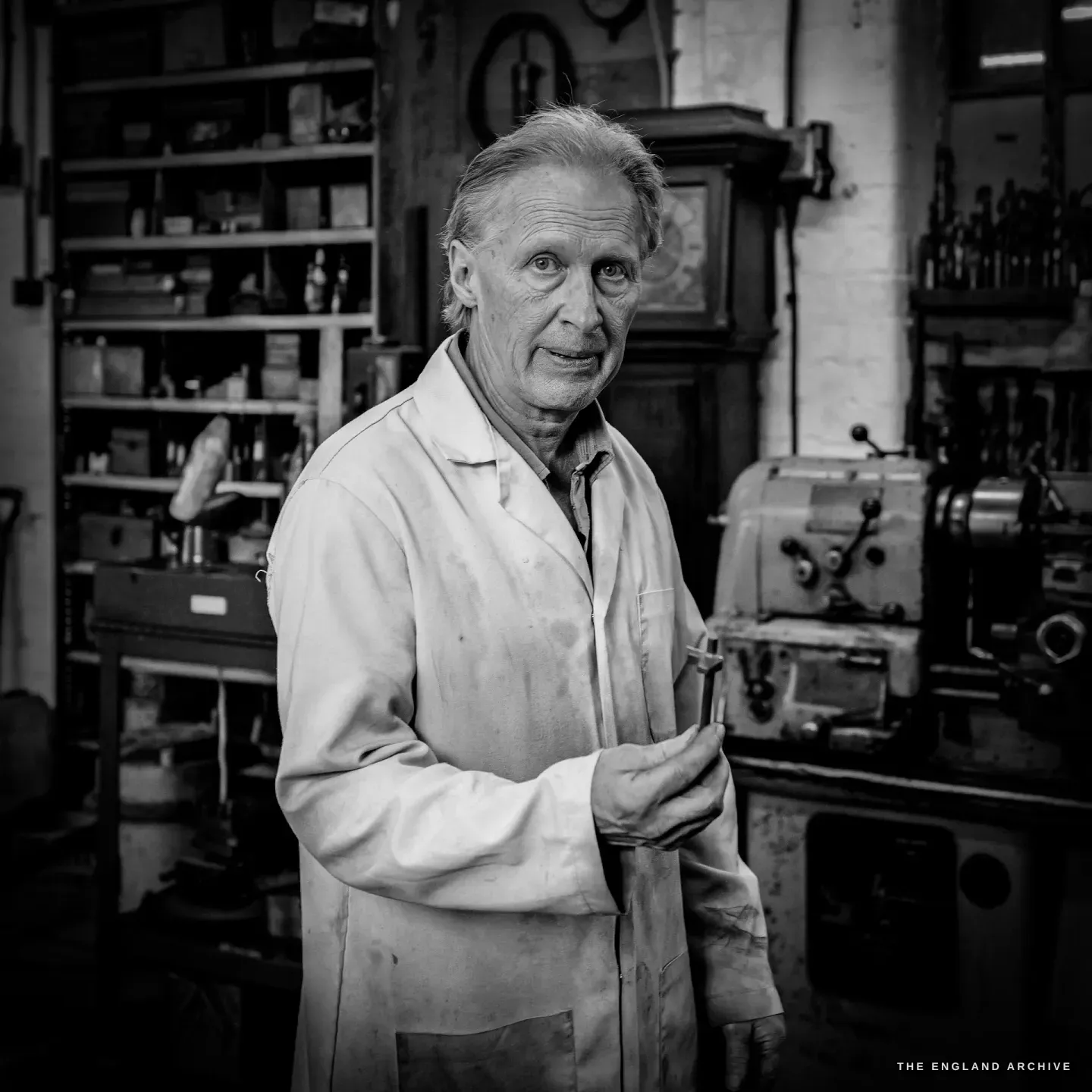 Haydn Hill in a white work coat faces the camera in the daylit workshop, a small tool in one hand, machines and shelves of parts behind him.