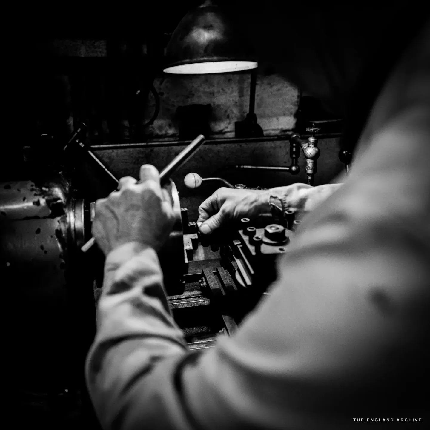 A view over Haydn Hill's shoulder as he works a part at the lathe, his forearm bright in the foreground and the machine and chuck beyond.