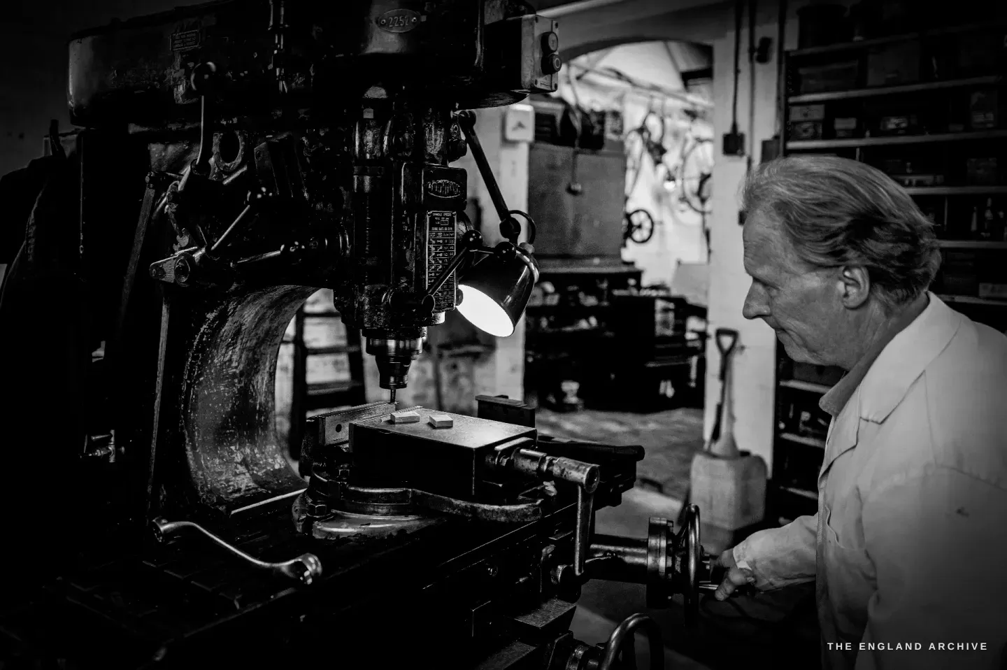 Haydn Hill works a large pillar drill lit by a small green-shaded lamp, turning the table handle, shelves and a bicycle behind him in the dark.