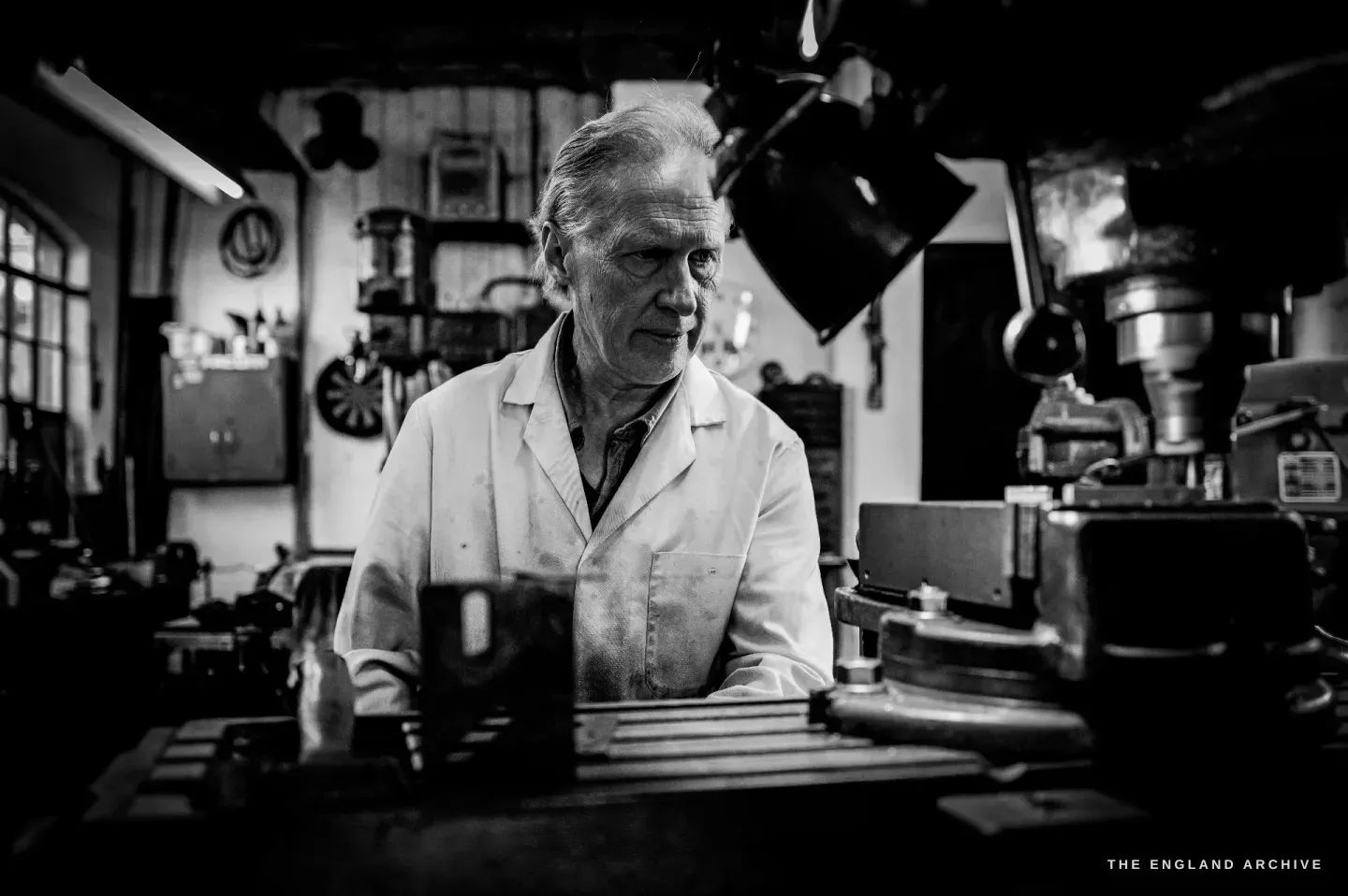 Haydn Hill stands behind a machine facing the camera under a strip light, the workshop and its shelves receding into shadow around him.
