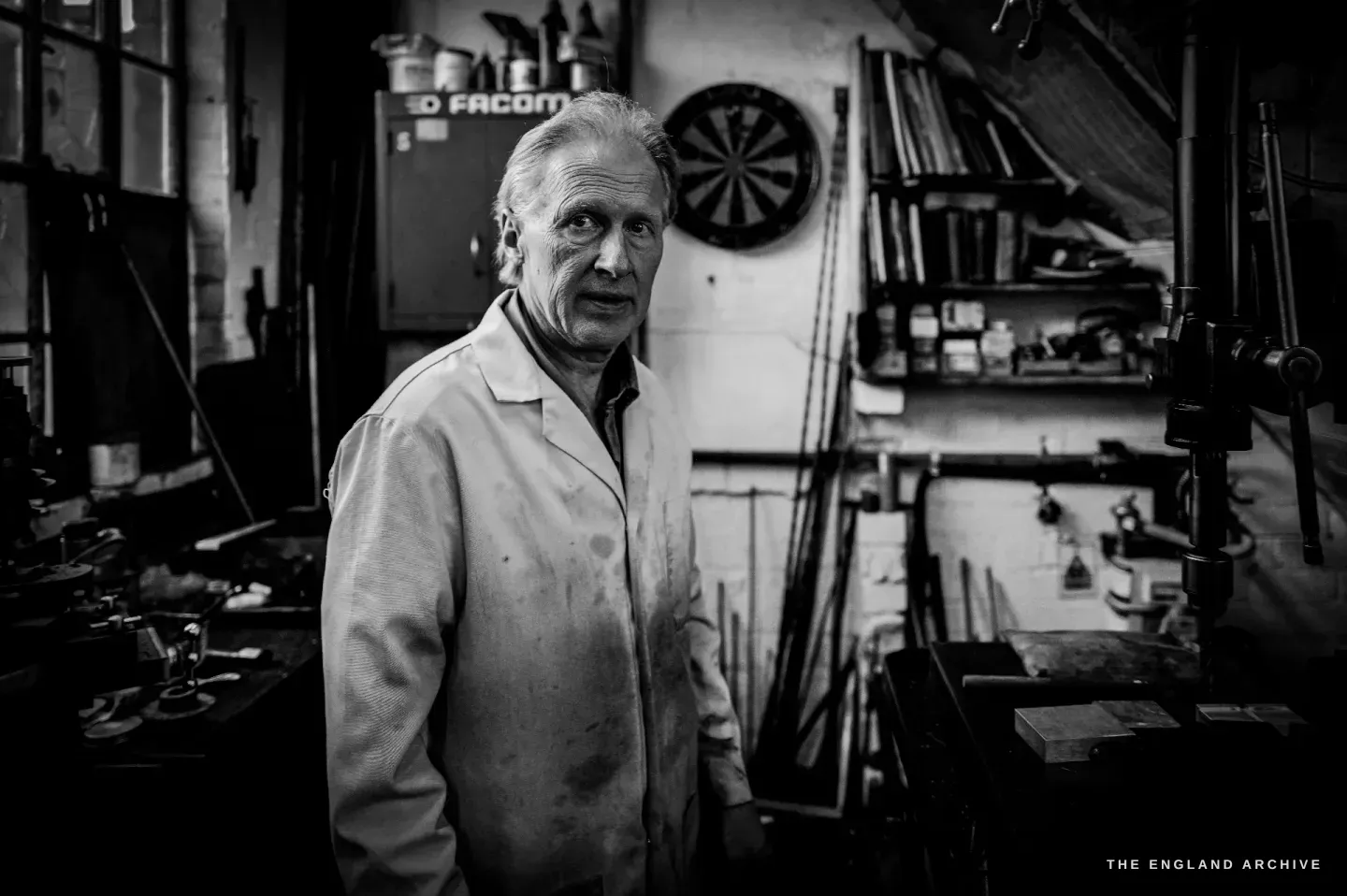 Haydn Hill stands three-quarters to the camera in his white coat, a dartboard and a red tool cabinet on the wall behind him, a machine in the foreground.
