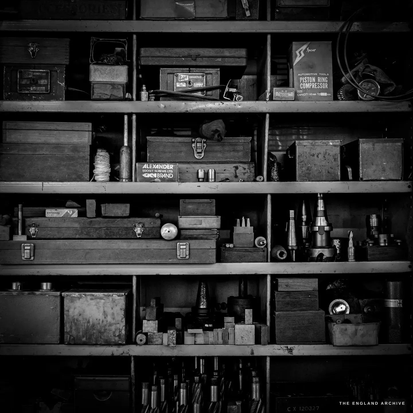 A tall steel shelf packed with old labelled tins and boxes, drill bits, gauges and a brass pressure gauge, a longcase clock just visible to the right.
