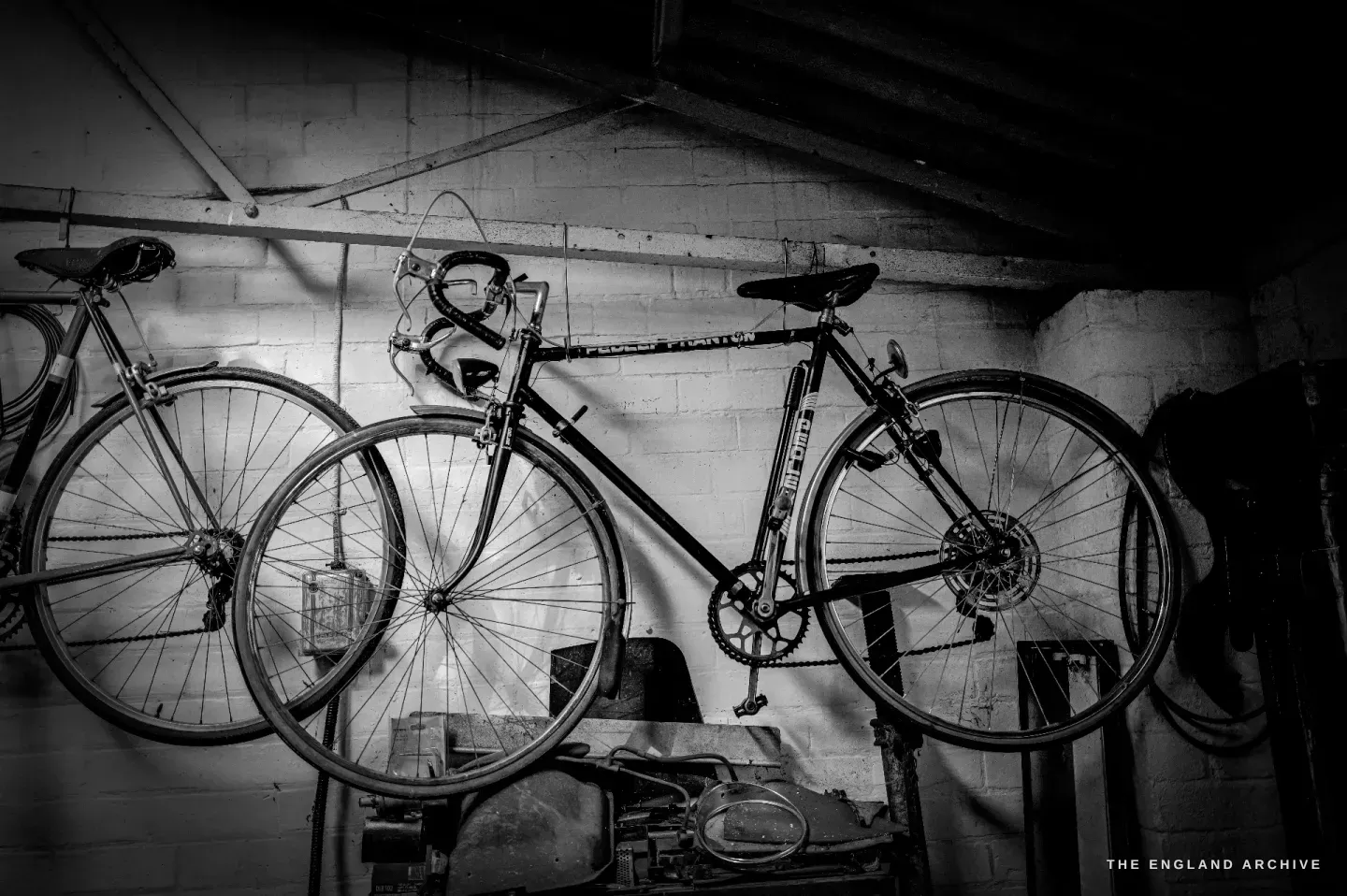 A road bicycle hung by its wheels on the whitewashed brick wall of the workshop, tools and boxes on the floor beneath it.