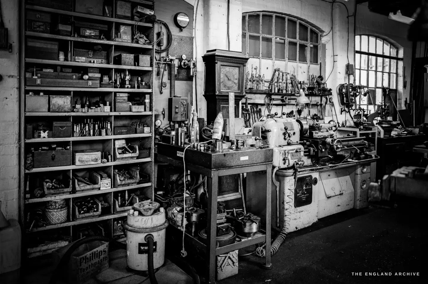 A wide view of the workshop: shelves of tins and tool boxes, a green centre lathe, a tall arched window and a longcase clock standing against the far wall.