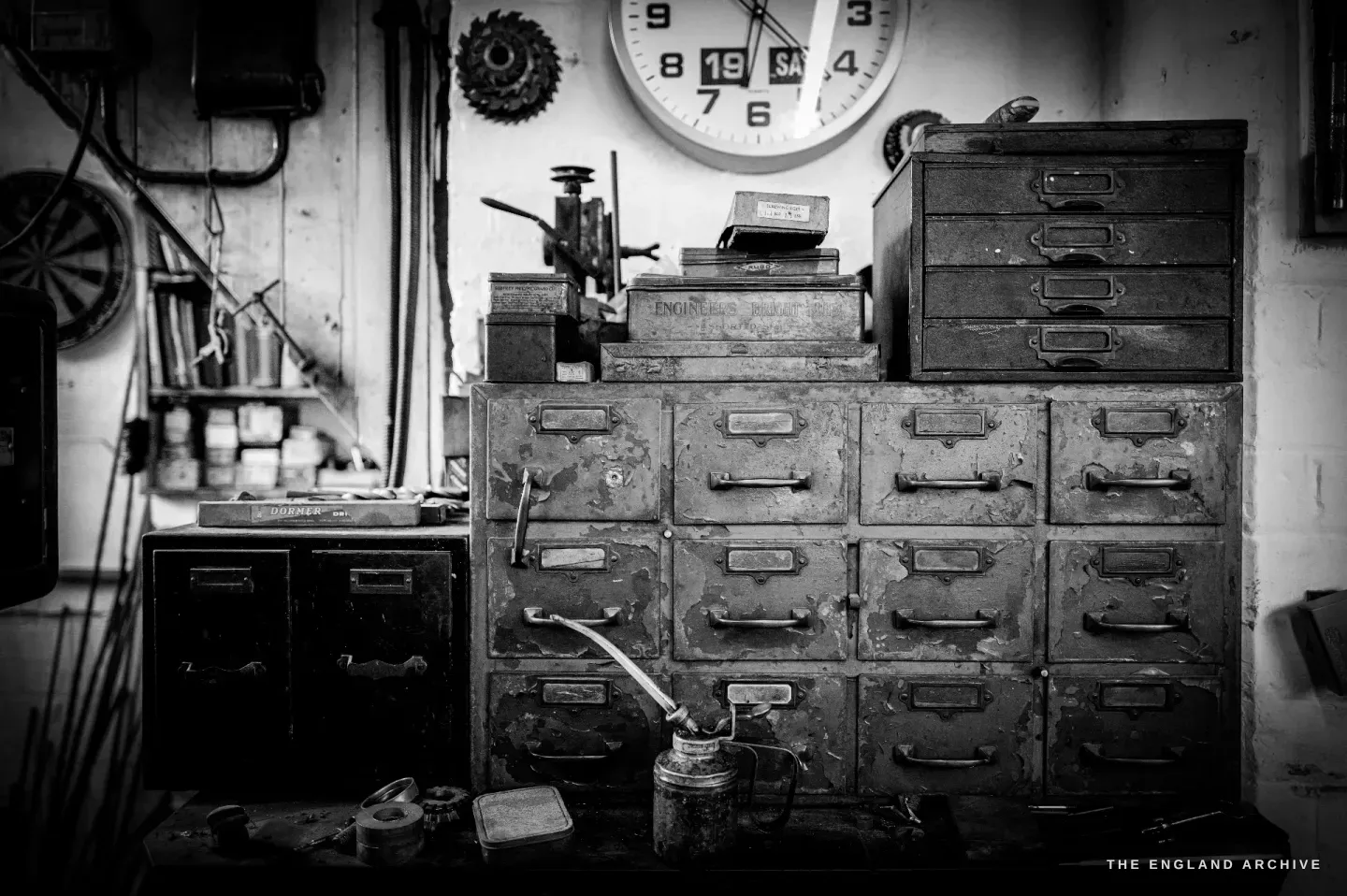 A corner of the workshop: a wall clock, a bank of little steel drawers and a drill press, tins and oil bottles crowded on the shelves.
