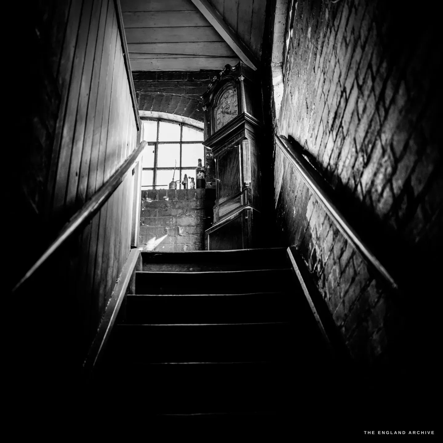 Looking down a dark, narrow staircase towards a landing lit by an arched window, a longcase clock standing at the foot of the stairs.