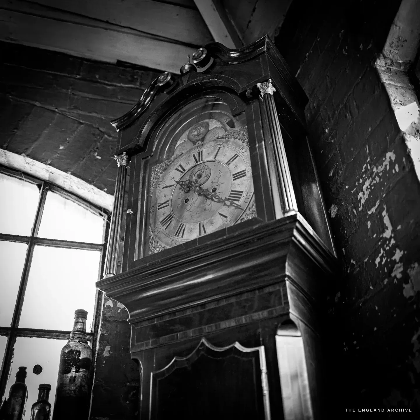A painted longcase clock standing against a brick wall beside a tall window, its arched dial catching the daylight, old bottles on the sill below.