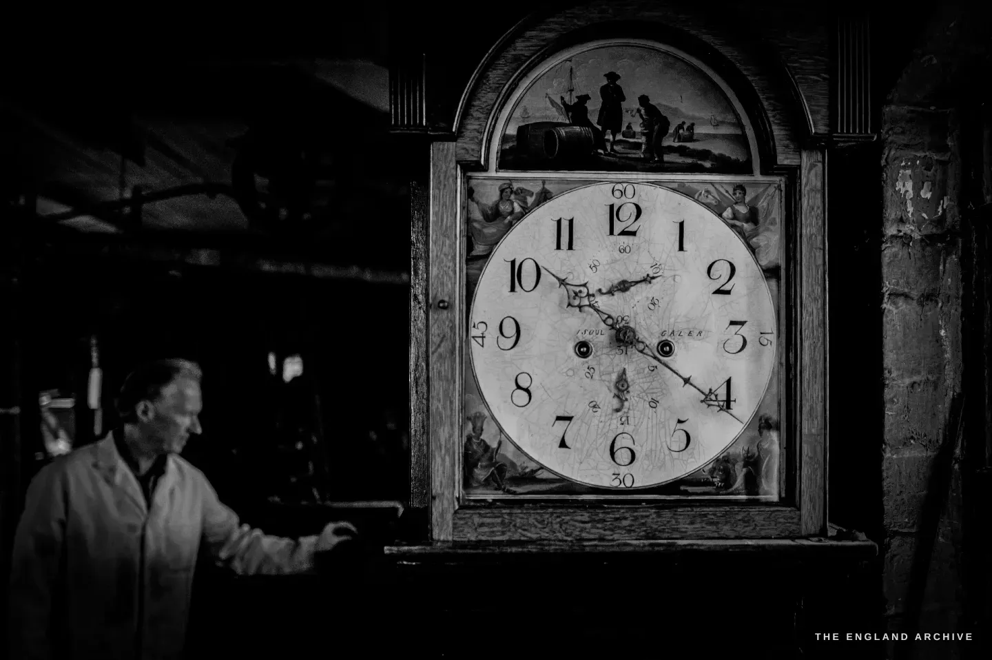 A finely painted longcase clock dial, its arch painted with small figures, Haydn Hill blurred in his white coat as he reaches to wind it.