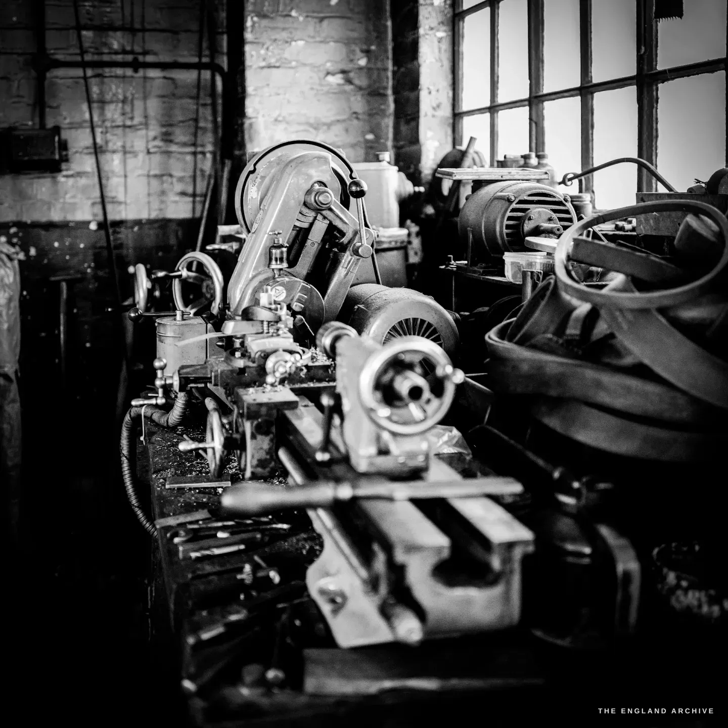 A long belt-driven lathe running down the middle of the workshop beneath a tall barred window, bar stock and hand tools laid along its bed.