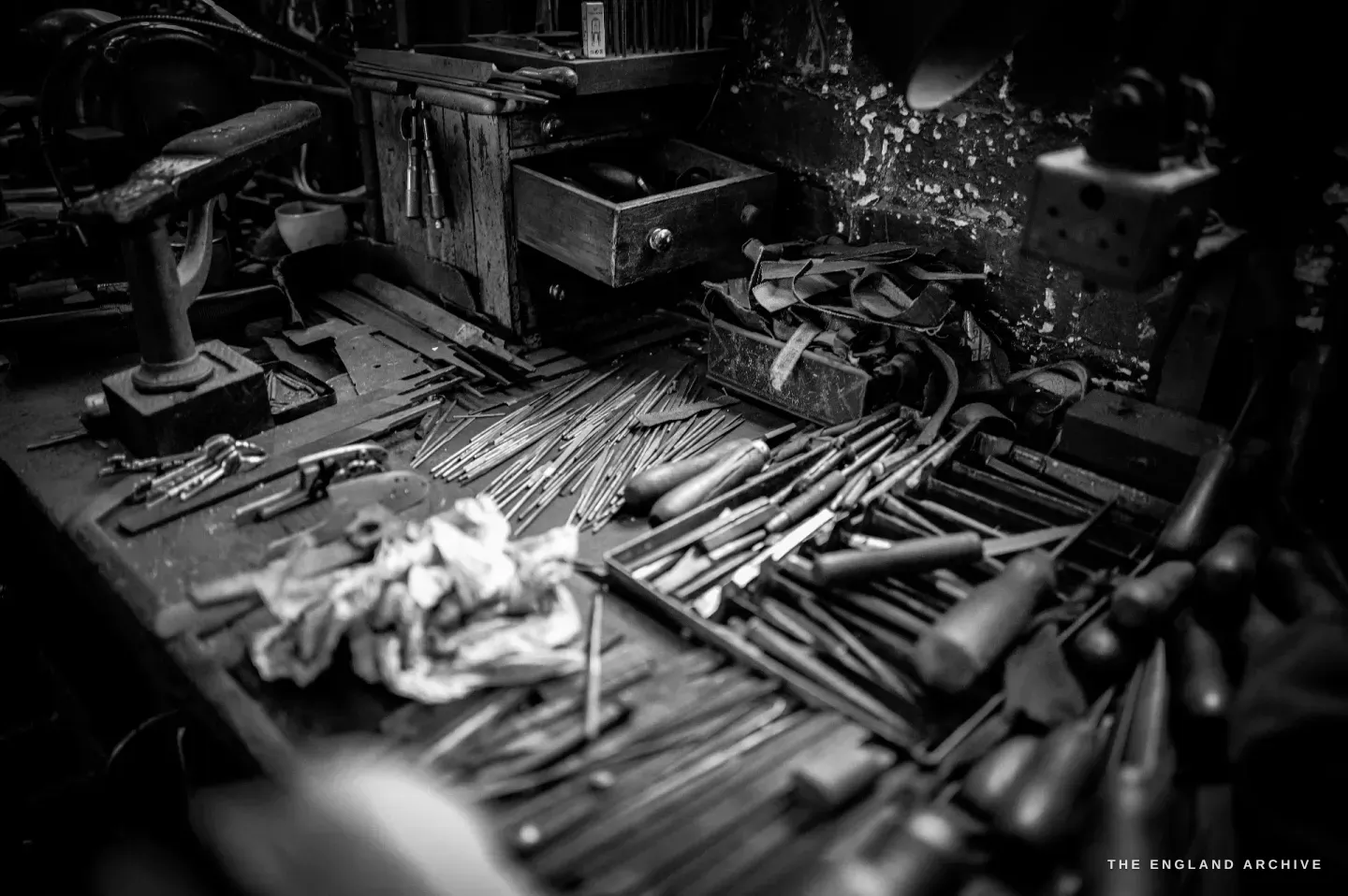 A bench spread with files, drills and hand tools laid across the wood, a drawered cabinet and a stitched leather pad behind.