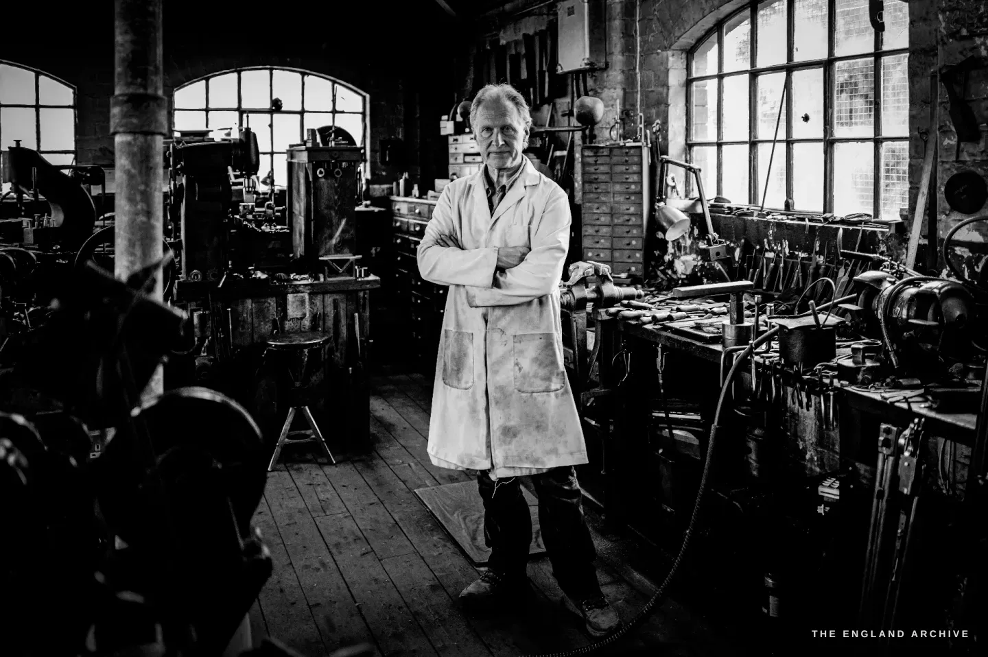 Haydn Hill stands with his arms folded in a white coat in the middle of the workshop, arched windows and benches of tools running away on either side.