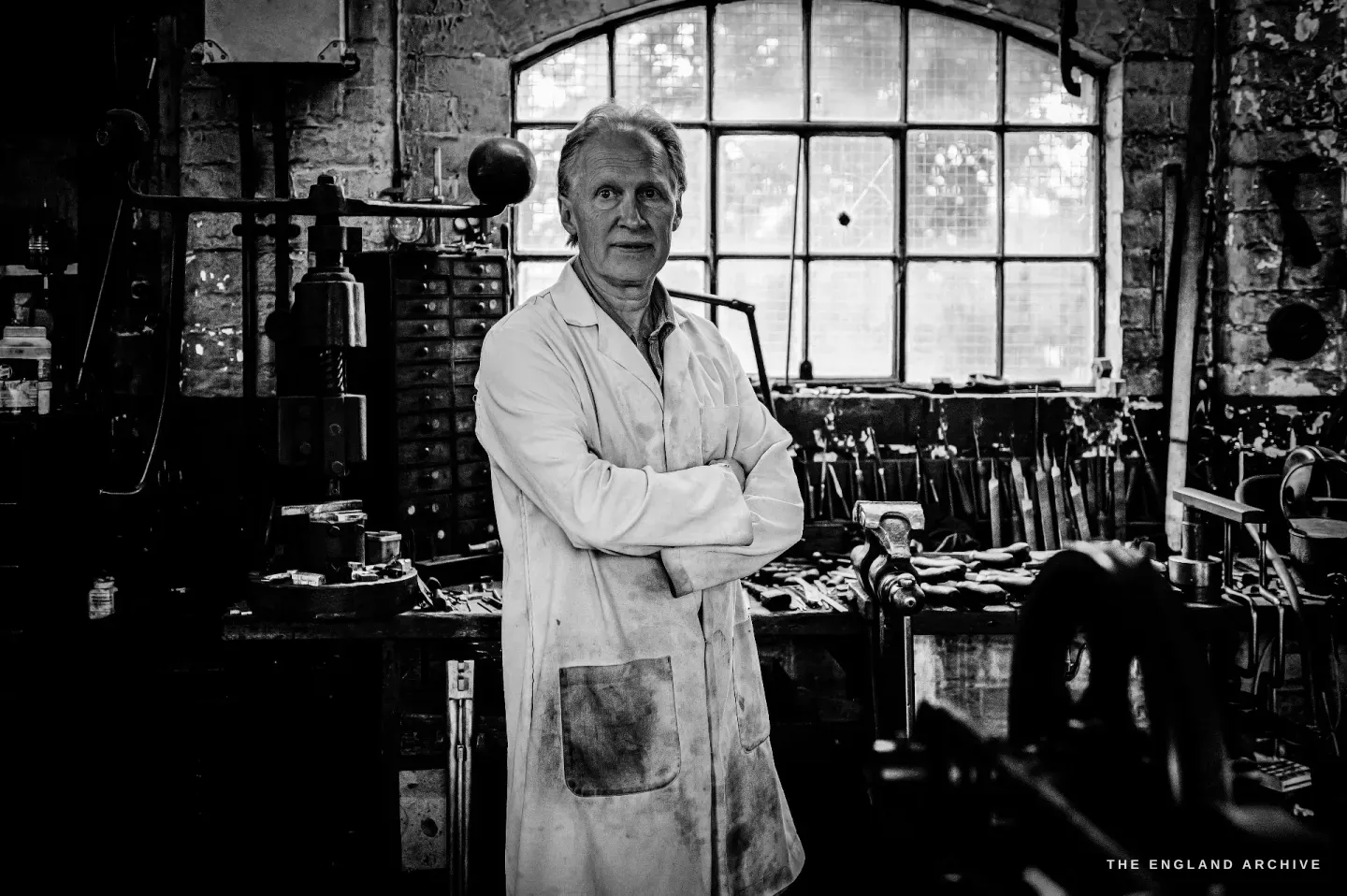 Haydn Hill stands, arms folded, in a stained white coat and apron in front of an arched workshop window, a bench crowded with tools beside him.