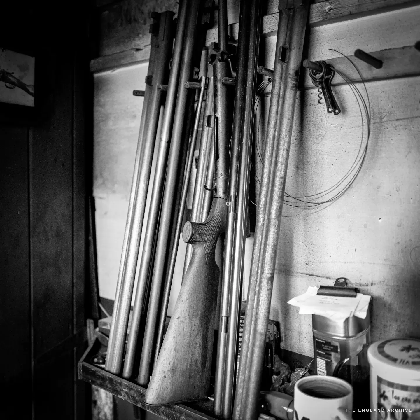A rack of gun barrels and an air rifle standing on end against a workshop wall, a mug and old tins on the shelf below.