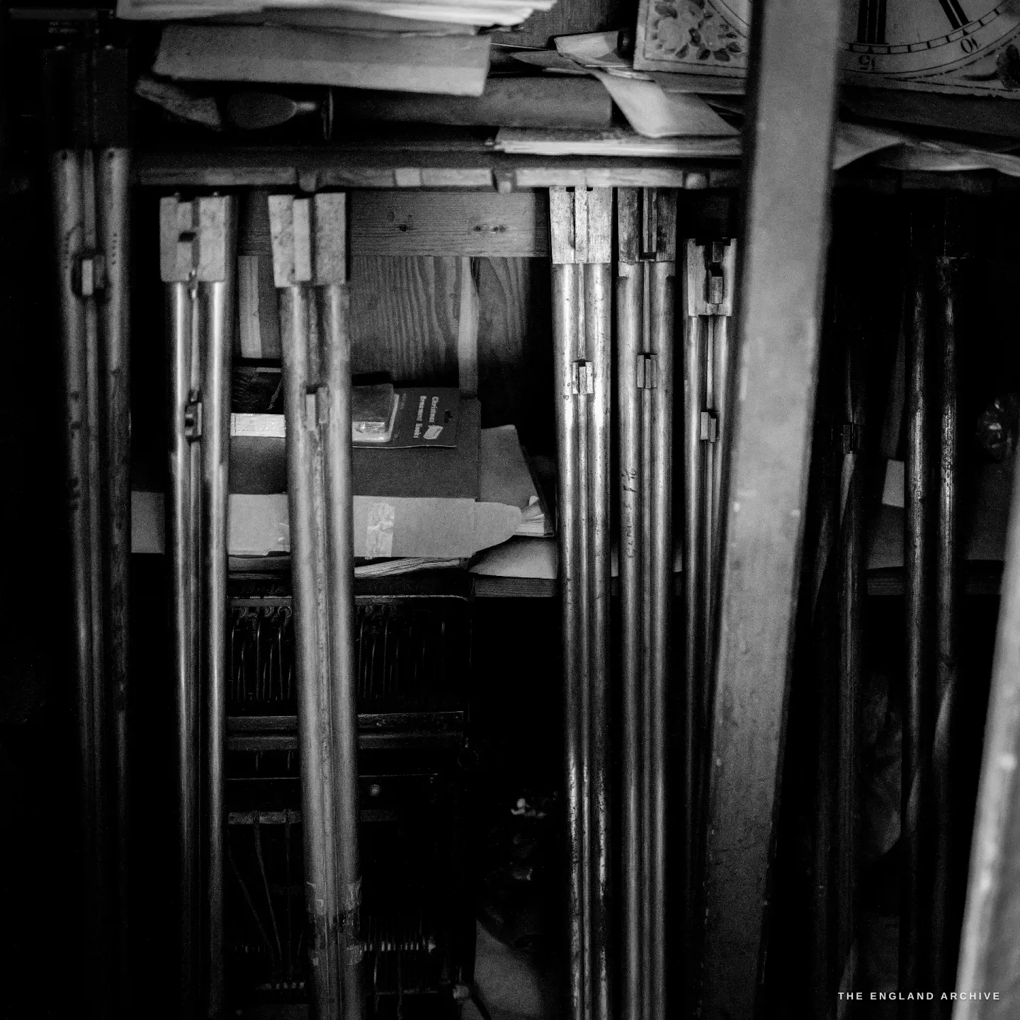 A dark storage nook stacked with blued double gun barrels standing on end, old papers and rolled drawings above.