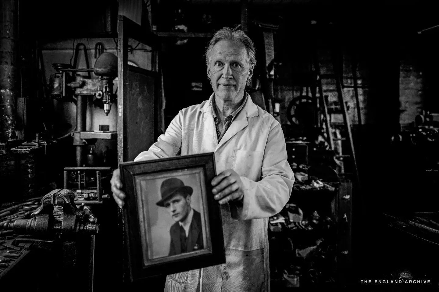 Haydn Hill holds up a small framed photograph of a young man in a hat, the machines of the workshop dark behind him.