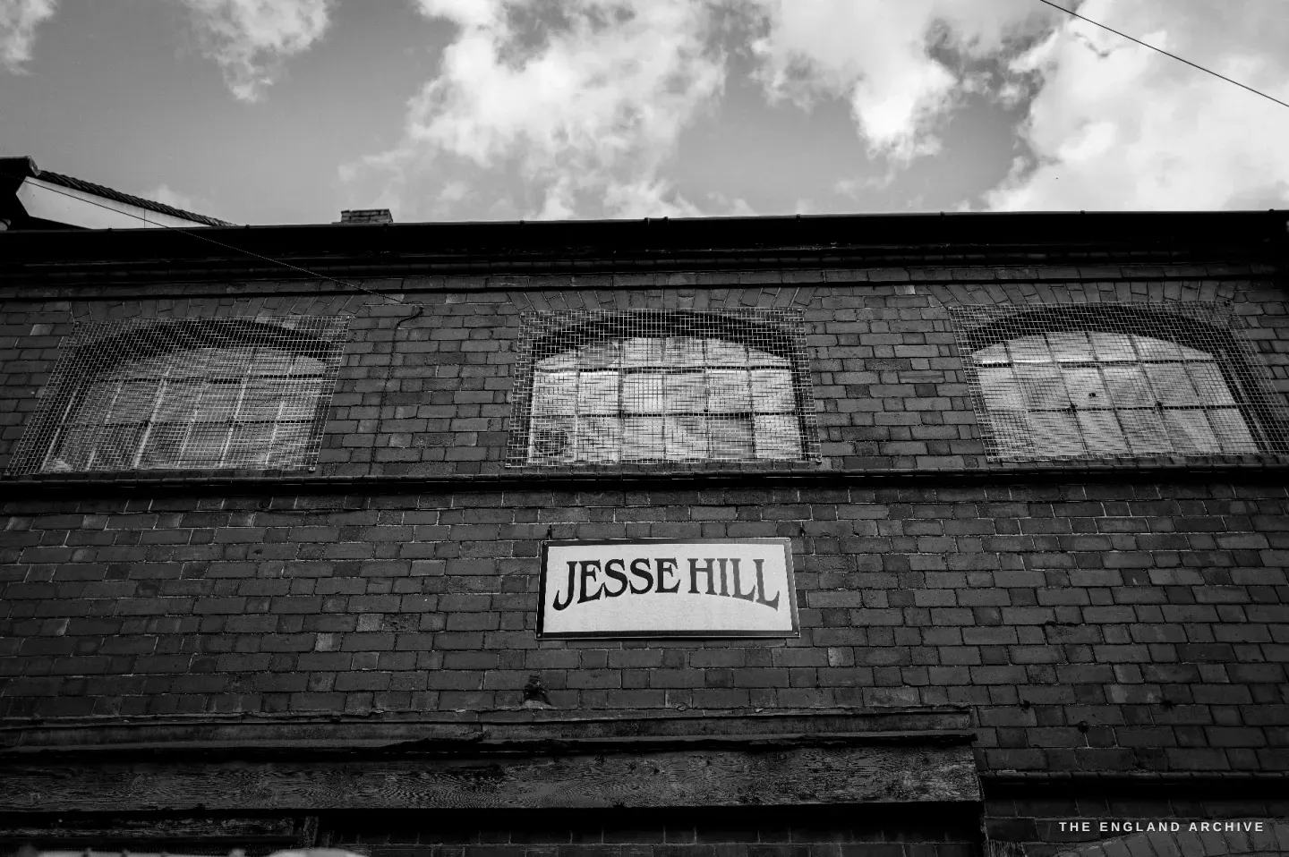 A painted JESSE HILL nameboard fixed high on the brick wall of the workshop, a row of arched windows below it and cloud behind.