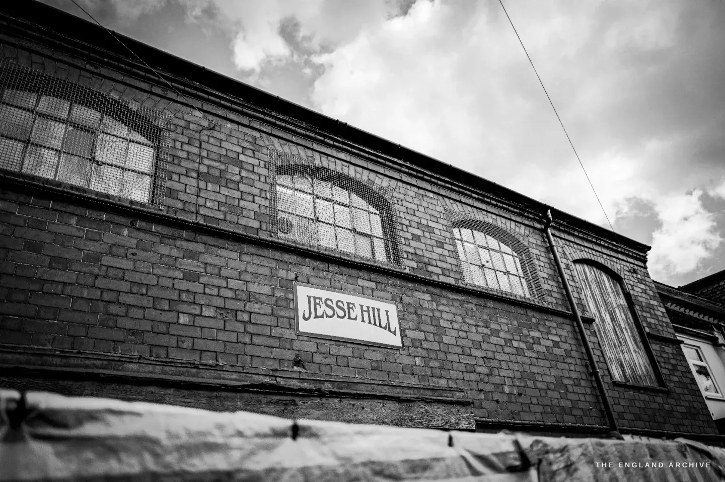 A low-angle view up the brick front of the workshop, the painted JESSE HILL board and the arched windows set against a bright, cloudy sky.
