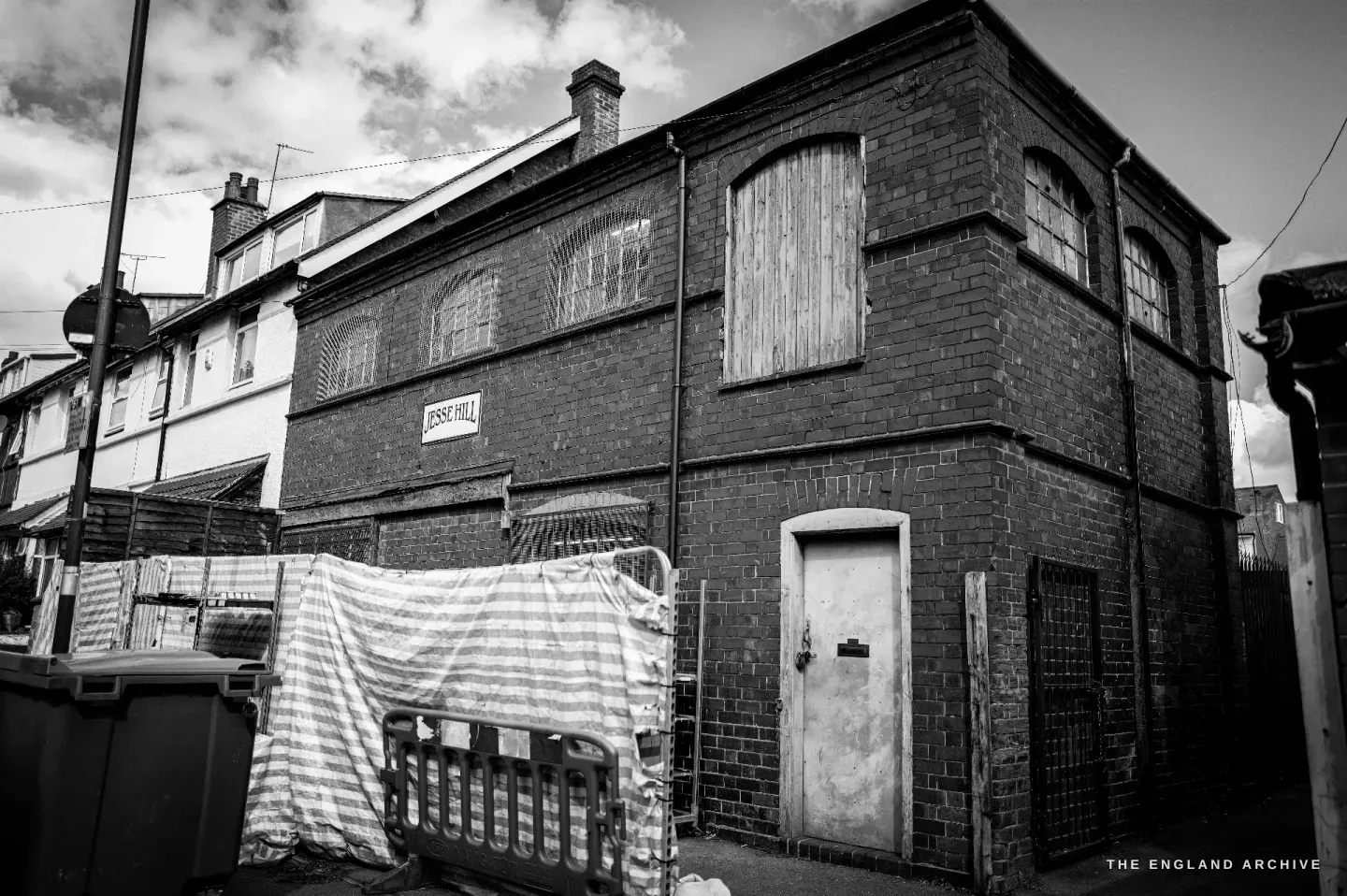 The Jesse Hill workshop seen from the street: a red-brick two-storey building with arched upper windows, a boarded-over loading door and a painted nameboard high on the wall.