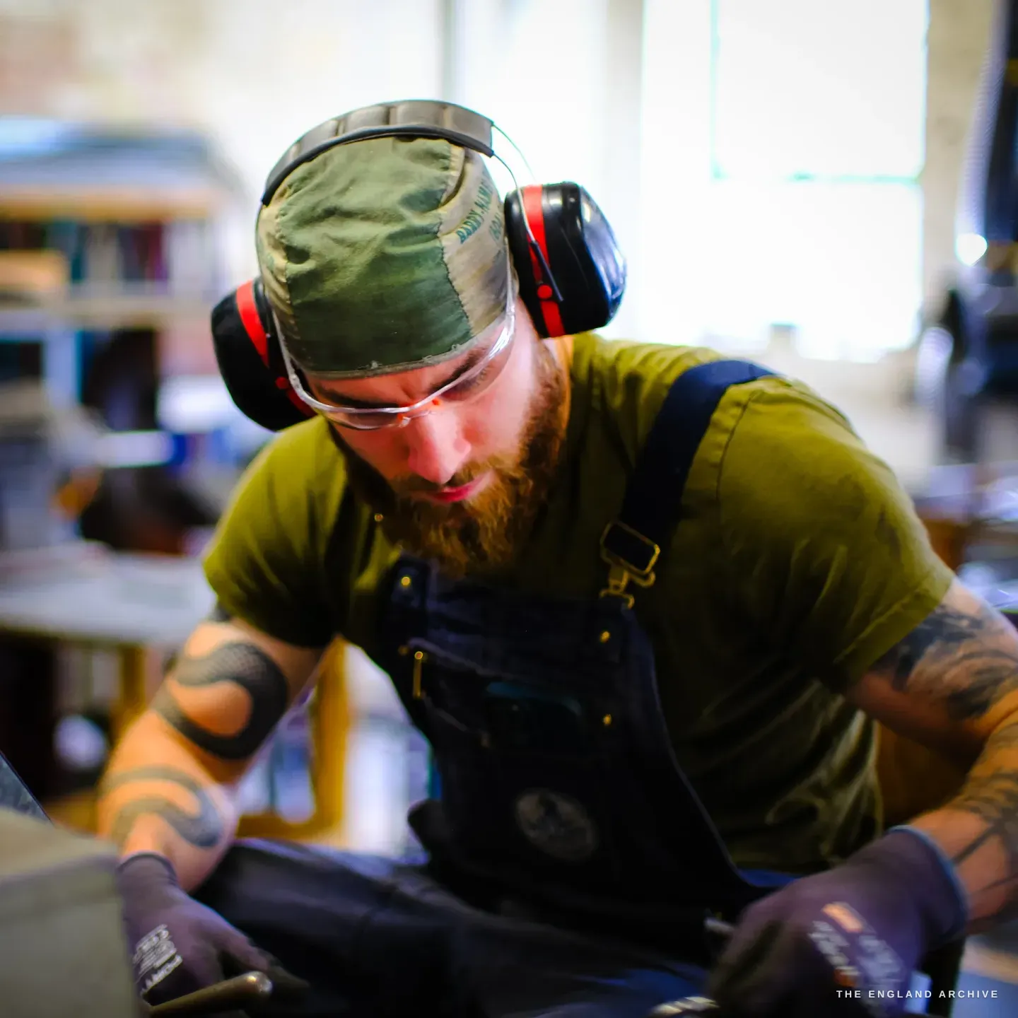 James bent close over his work, cap and ear defenders on, safety glasses down, beard and a tattooed arm catching the light.