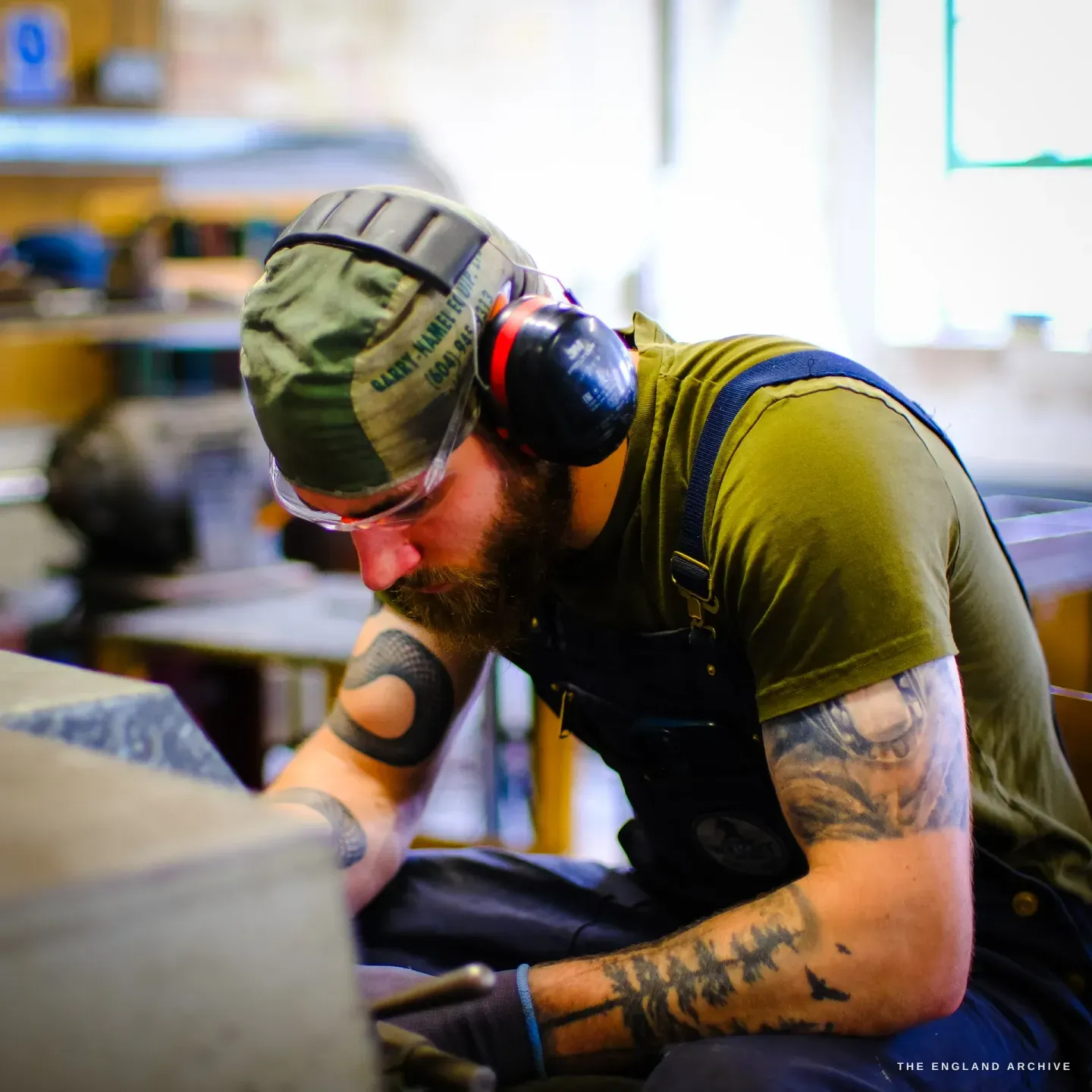 James in profile bent over the machine, working a blade, the bright workshop windows behind him.