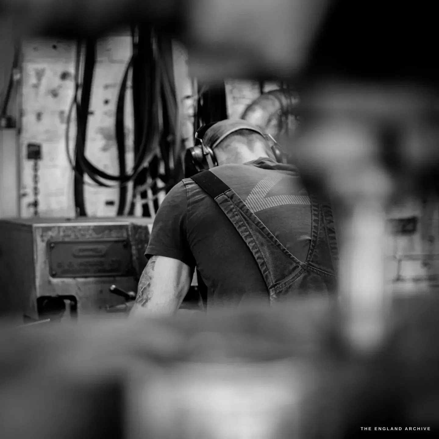 James seen from behind through racked blades, ear defenders on, at his machine, a maker’s plate and cabling on the wall beyond.