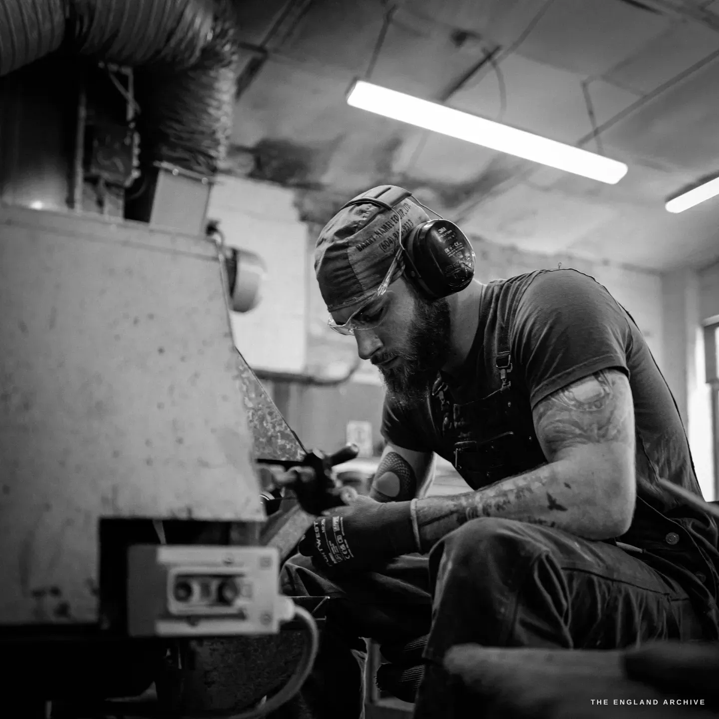 James seated at his grinding machine under the fluorescent light, gloved hands holding a blade to the wheel, the machine controls at its base.