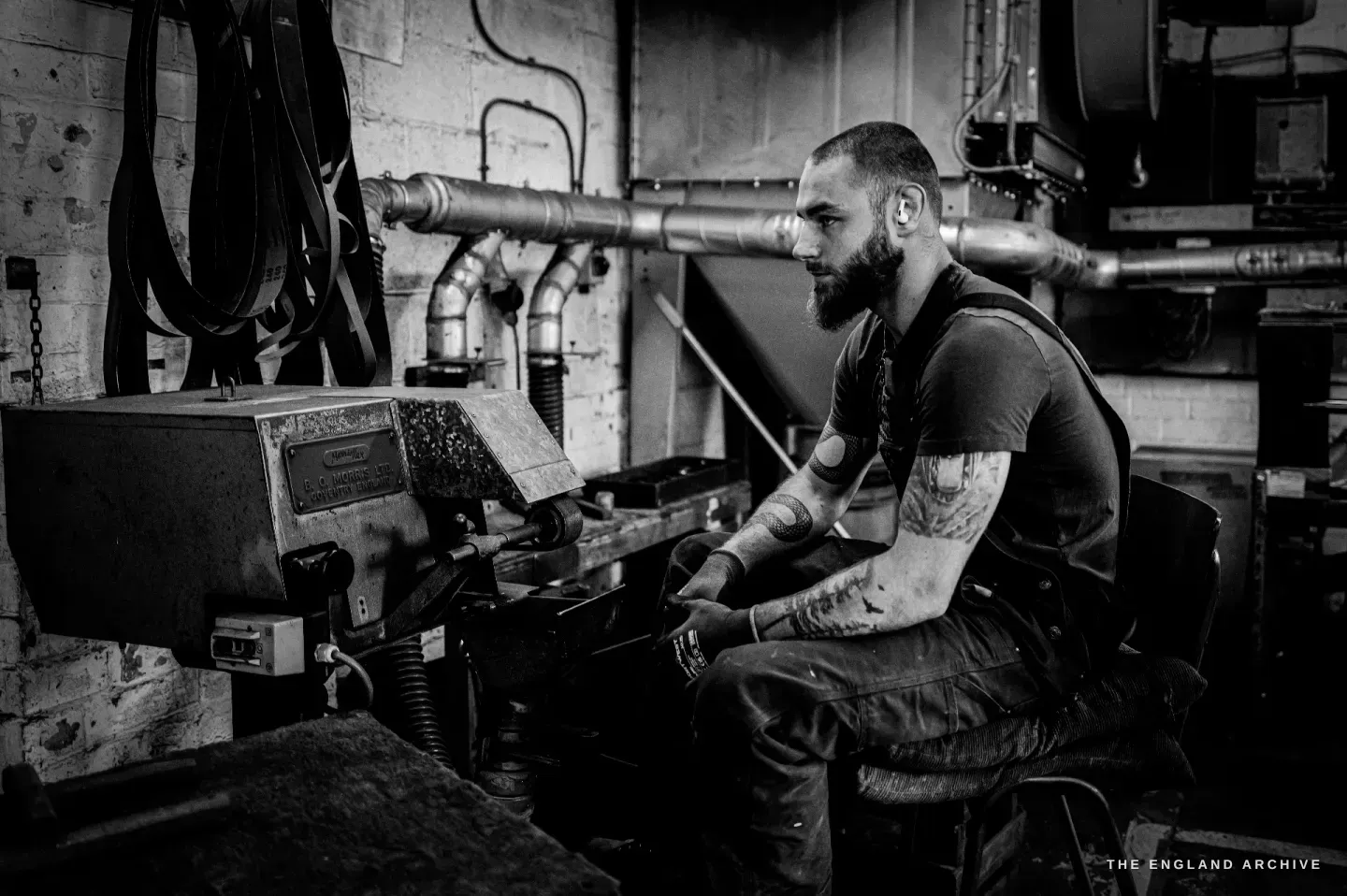 James seated in profile at his long cast-iron grinding machine, the polished extraction pipe running overhead, working a blade.