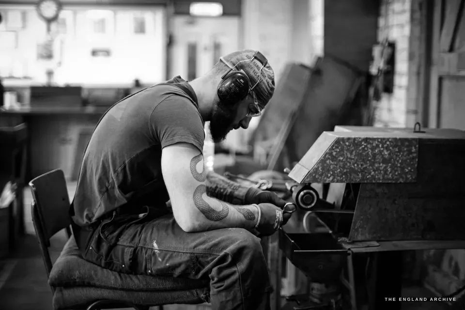 James Morton seated at his grinding machine in a cap and ear defenders, tattooed arms, bent to the work, the workshop behind him.