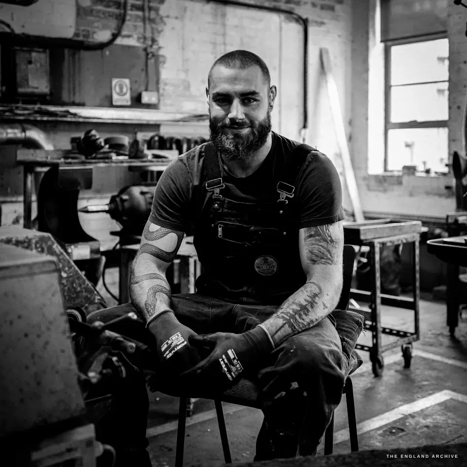 James Morton seated at his machine, shaved head and full beard, tattooed arms, gloved hands resting, looking off-camera with a slight smile, the bright workshop behind him.