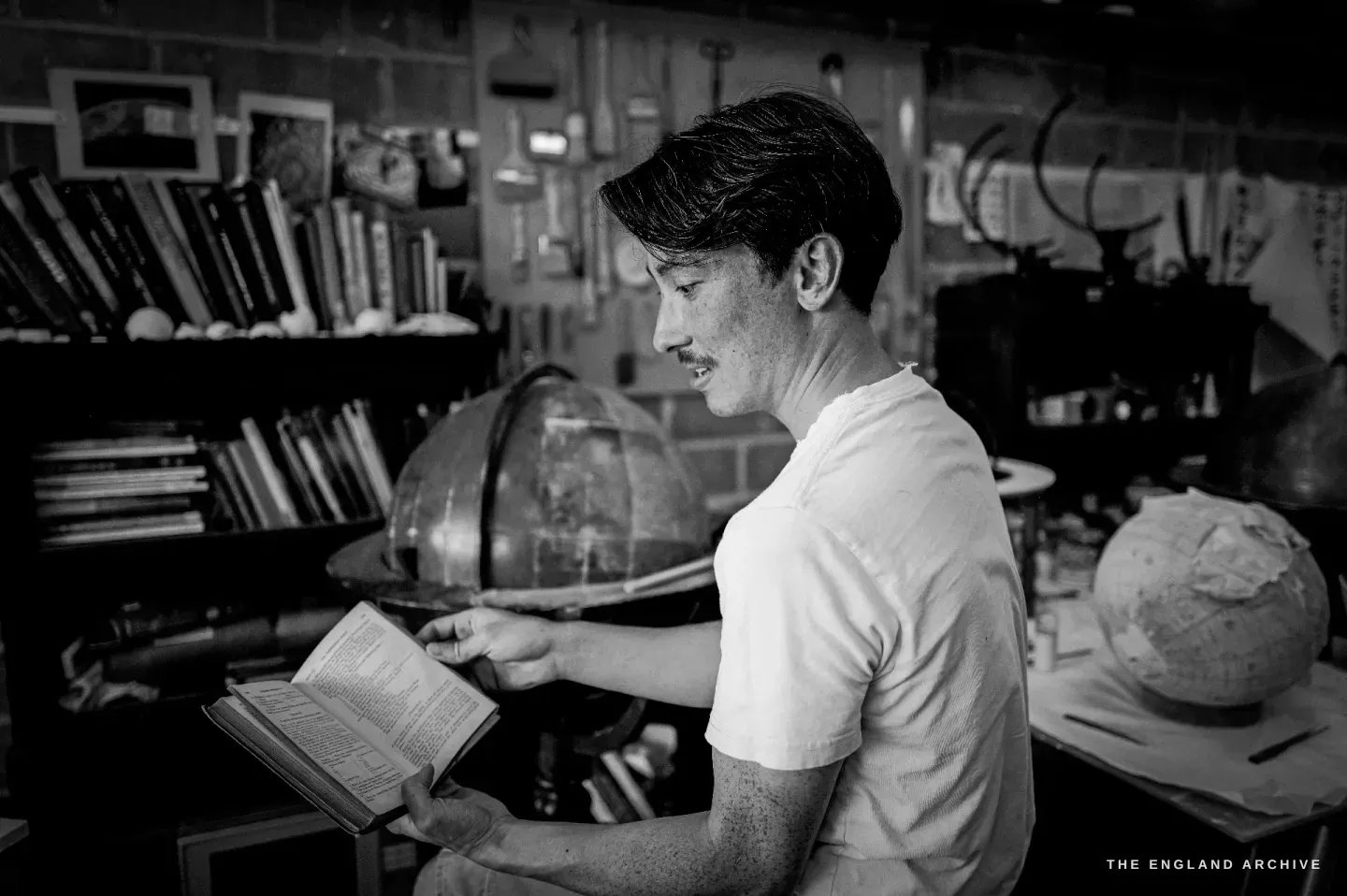 Jonathan seated reading a small old book, smiling, a globe and a bare globe core on the shelves around him.