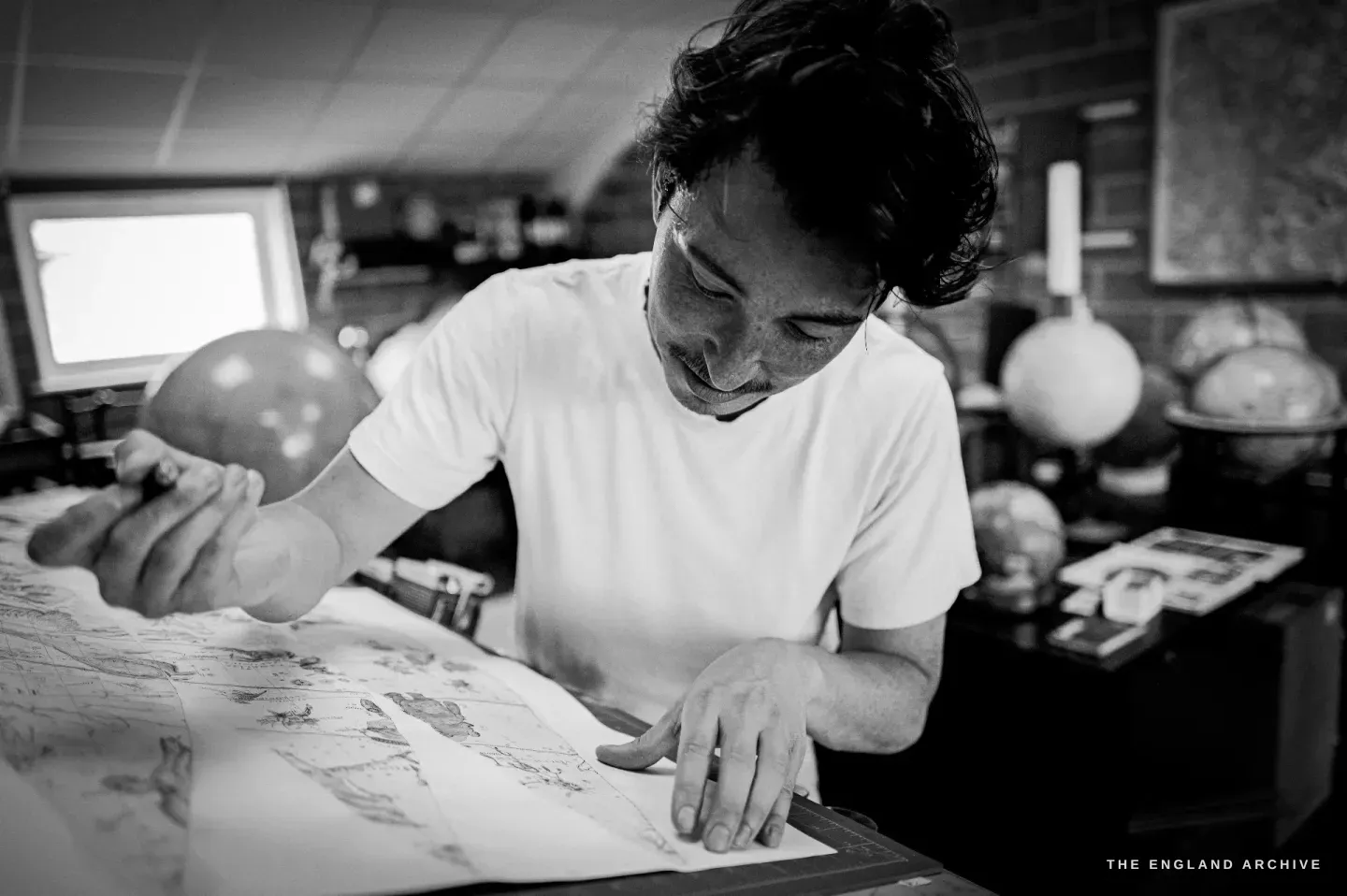 Jonathan smiling, holding a small globe in one hand and pointing at printed gore sheets with the other, finished globes on the shelves behind.