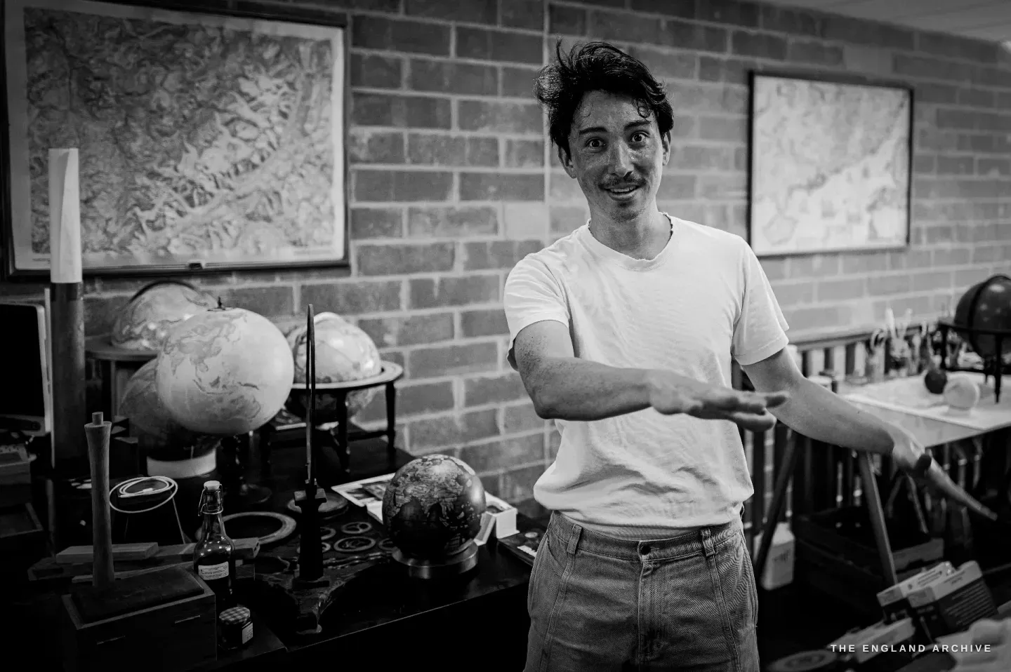 Jonathan standing in the workshop, gesturing as he talks, framed maps and a shelf of globes behind him.