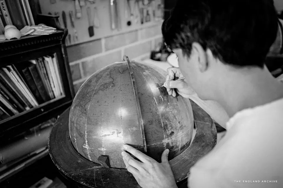 Jonathan leaning over a large globe, steadying it with one hand and working the surface with a tool, a wall of tools behind.