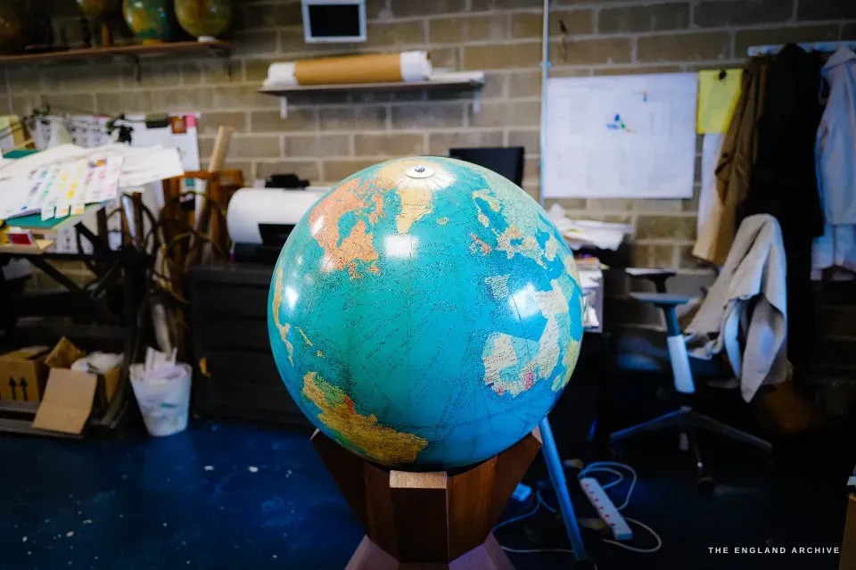 A newly made terrestrial globe in full colour - deep blue oceans, hand-tinted continents - on a turned wooden stand in the workshop.