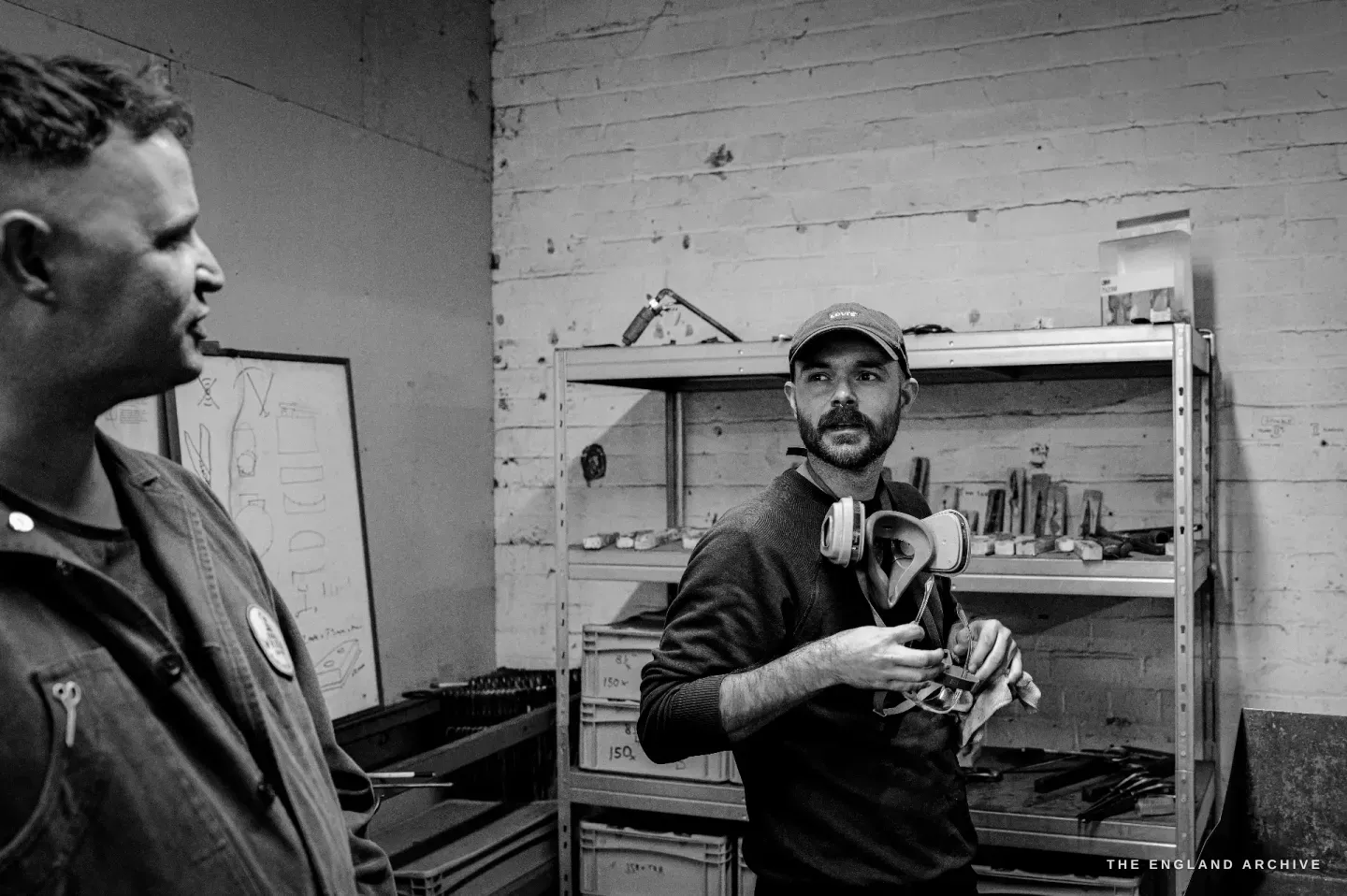 Jonathan in conversation with another maker, a respirator hanging at his neck and a cloth in his hands, racked stock behind.