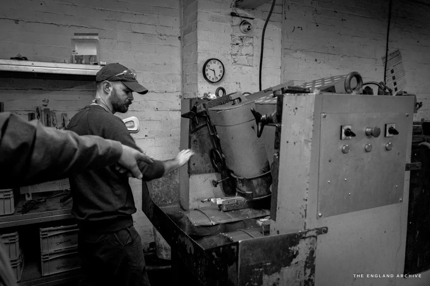Jonathan beside the open grinding machine, its hood swung up, a colleague’s hand reaching across to the work, a clock on the brick wall behind.